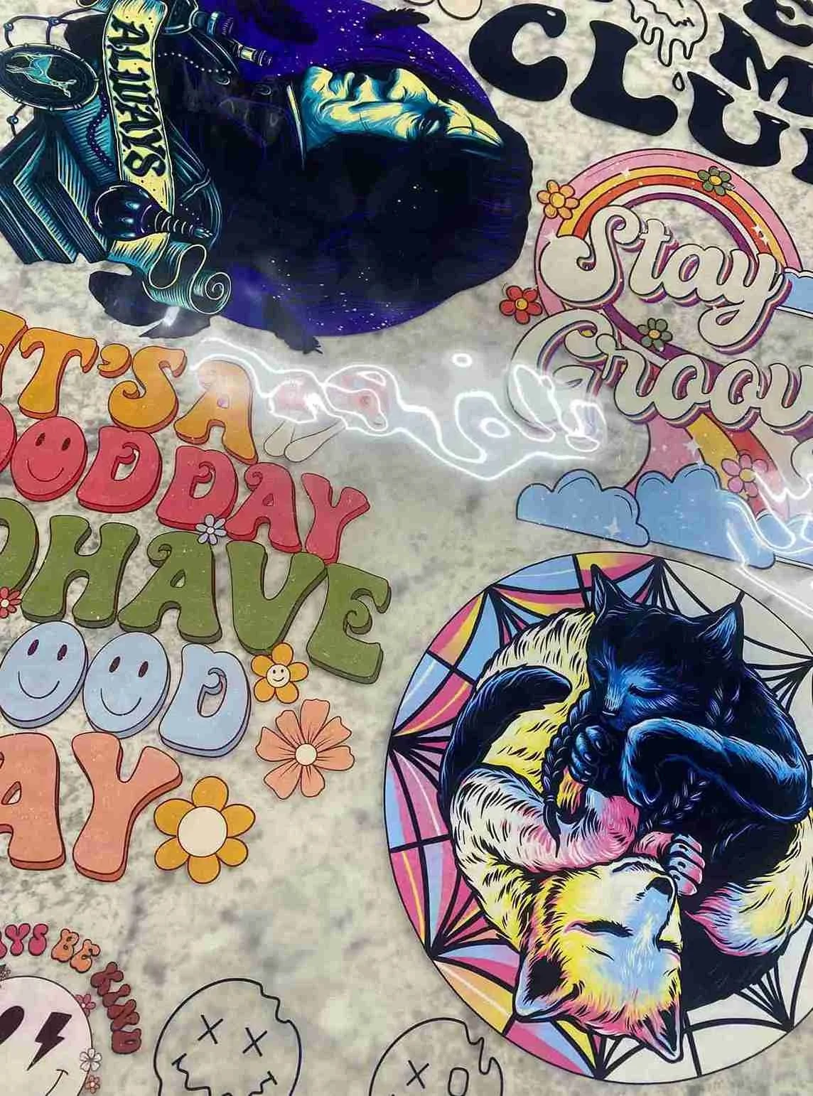 Colorful stickers on a surface, including a black dog with a blue glowing outline, a black cat curled up inside a rainbow-colored geometric shape, and positive messages like 'Stay Trippy' and 'It's a Good Day' surrounded by flowers and clouds.