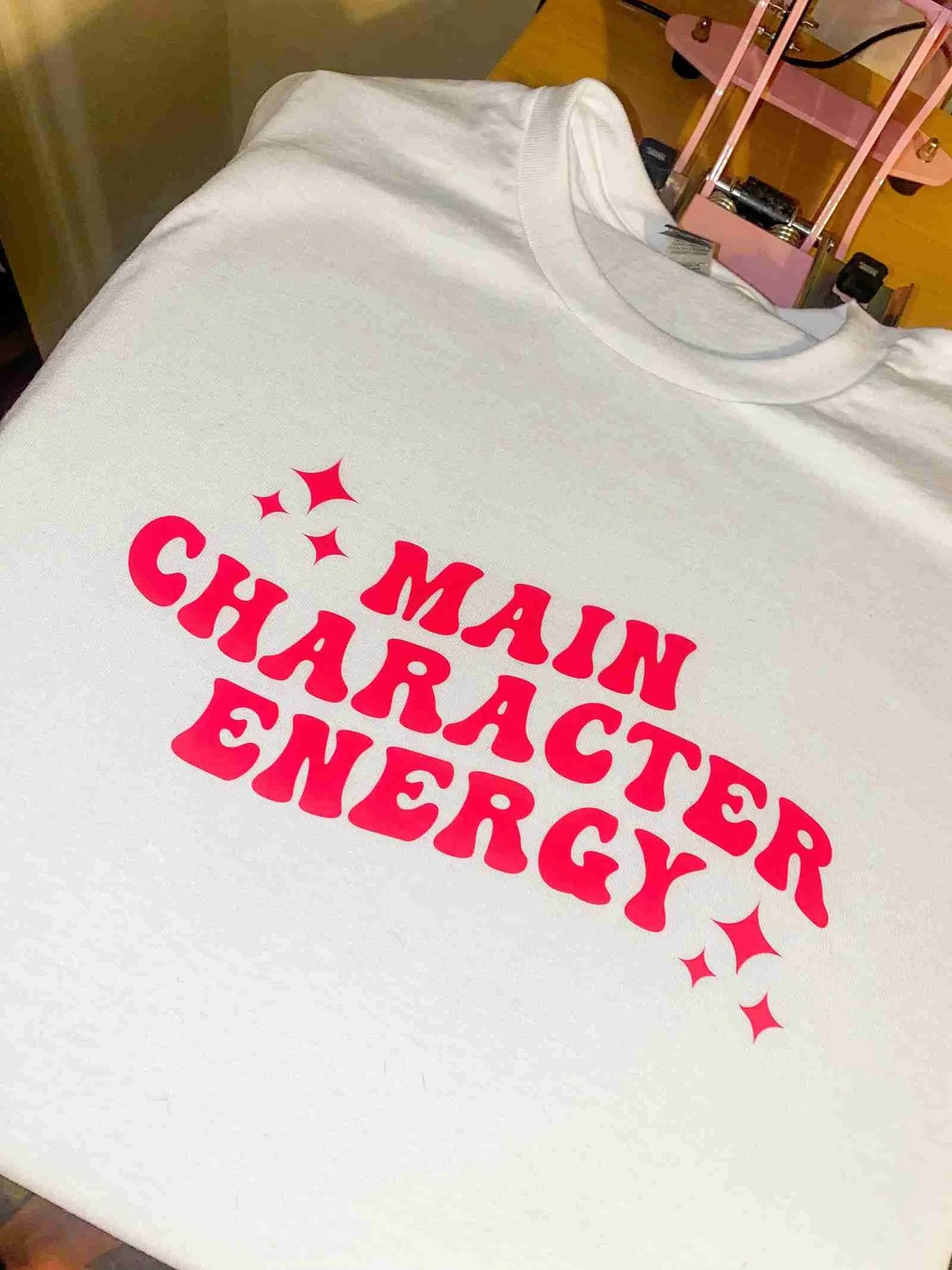 White t-shirt with bright pink text that reads 'Main Character Energy' surrounded by pink stars.
