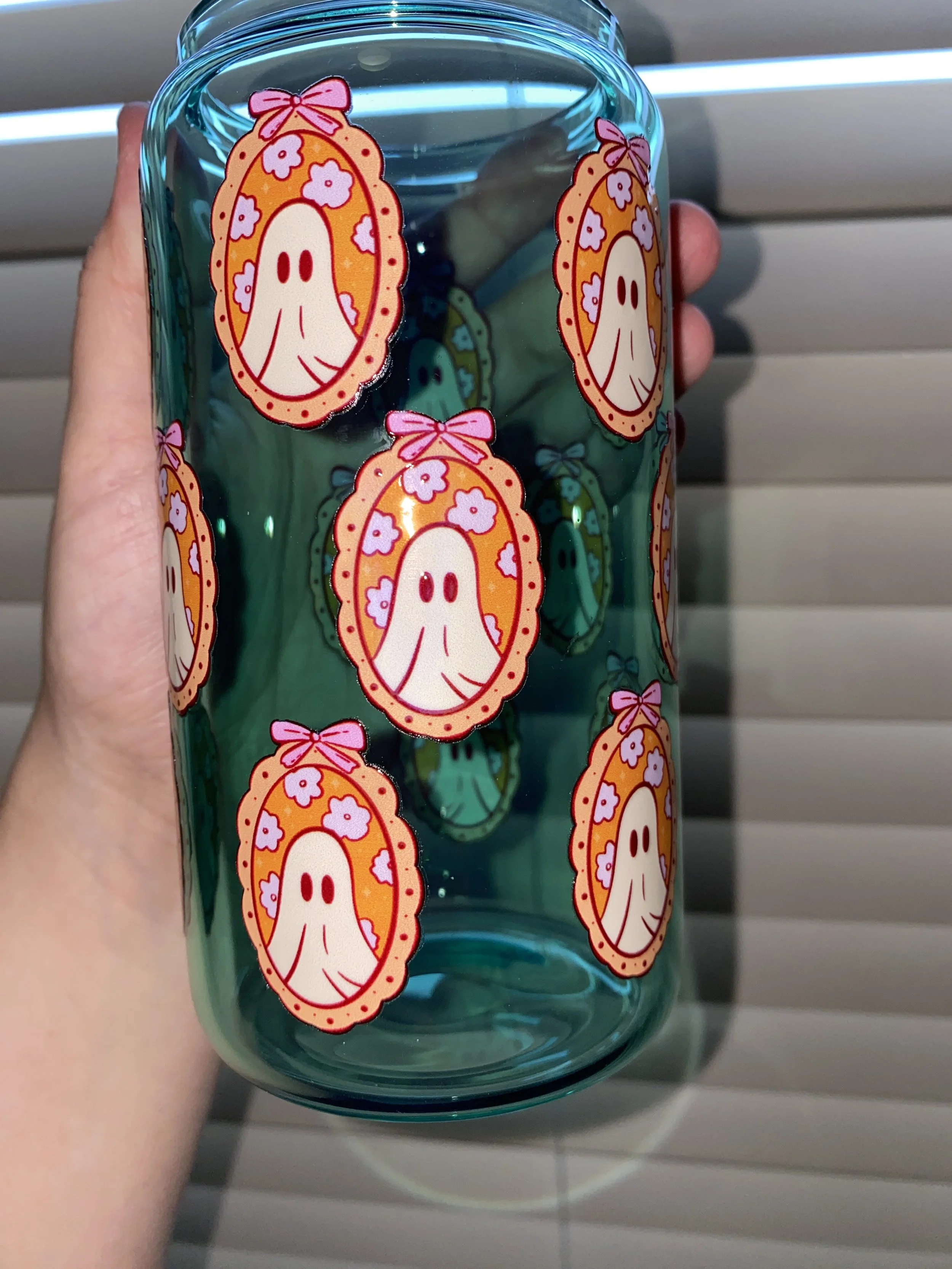 Hand holding a blue glass tumbler decorated with stickers of a ghost inside an oval frame with pink flowers and a pink bow.
