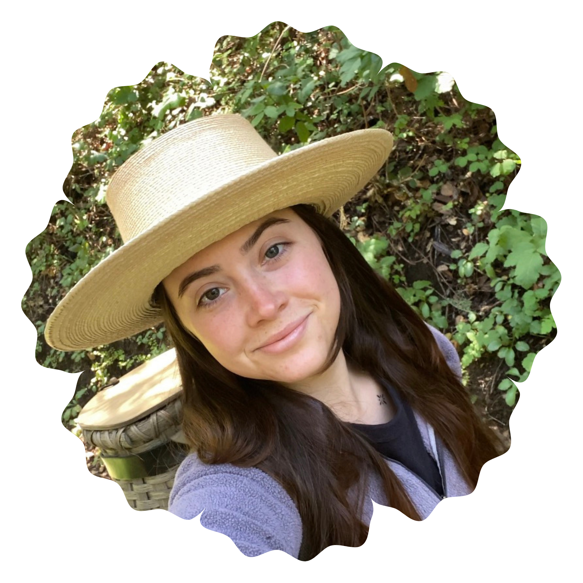 A woman with long brown hair and fair skin lying outdoors on green foliage, wearing a large straw hat and a purple fuzzy sweater.