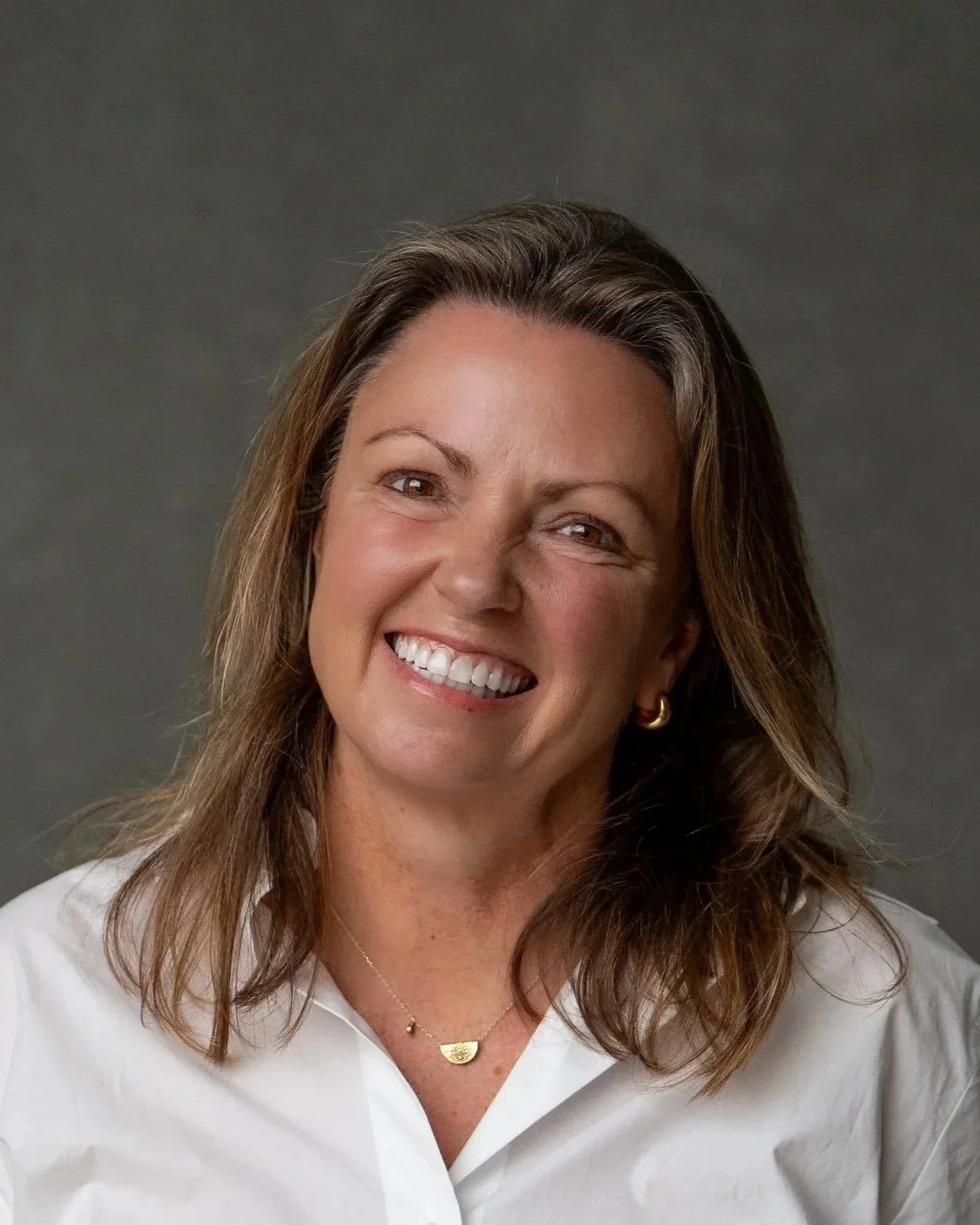 Jane Pincott, Naturopath and Nutritionist with shoulder-length brown hair, wearing a white shirt, gold earrings, and a gold necklace, against a neutral gray background.
