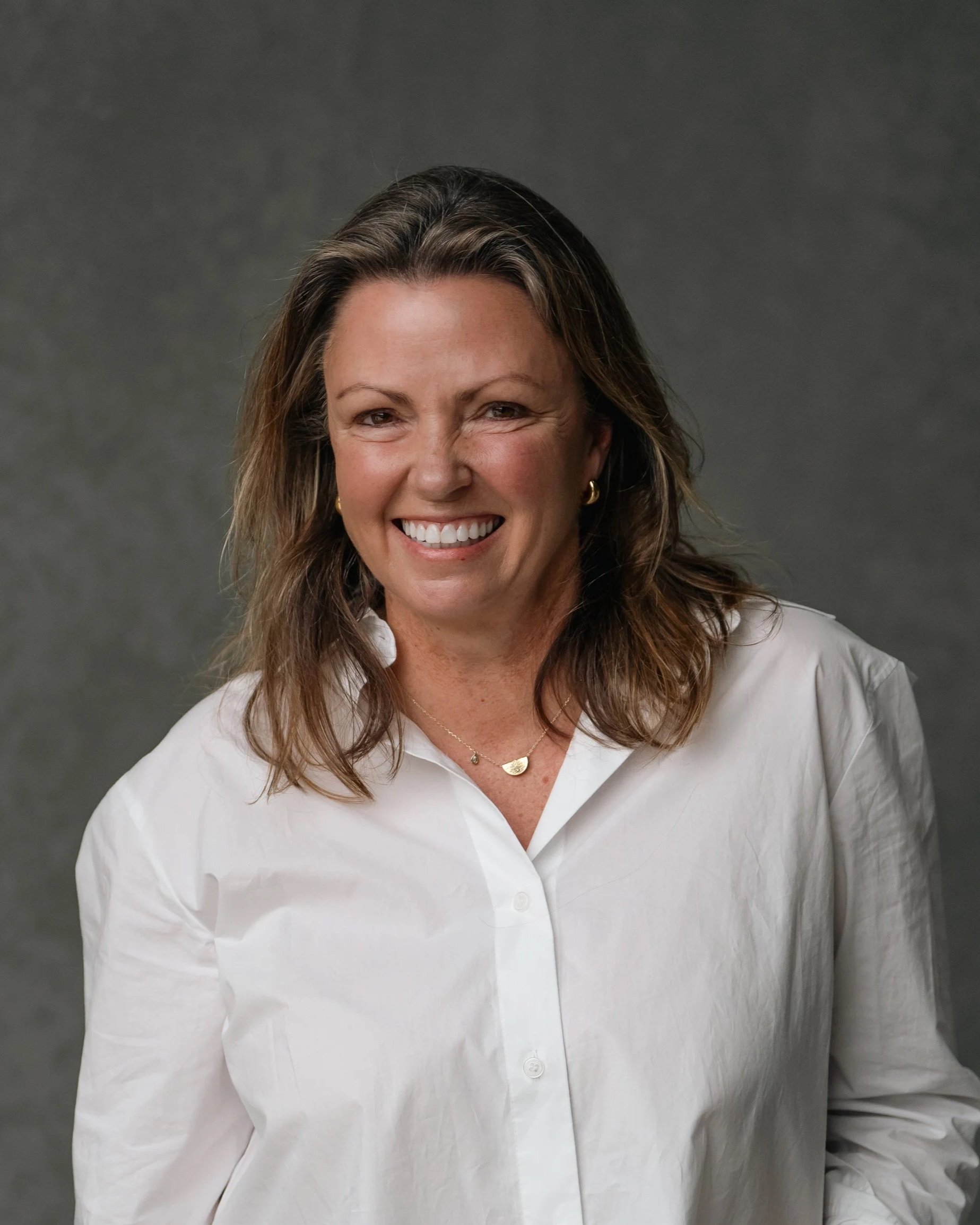 Close-up of Jane Pincott with shoulder-length brown hair, wearing a white shirt, gold hoop earrings, and a gold necklace, standing against a dark gray background.