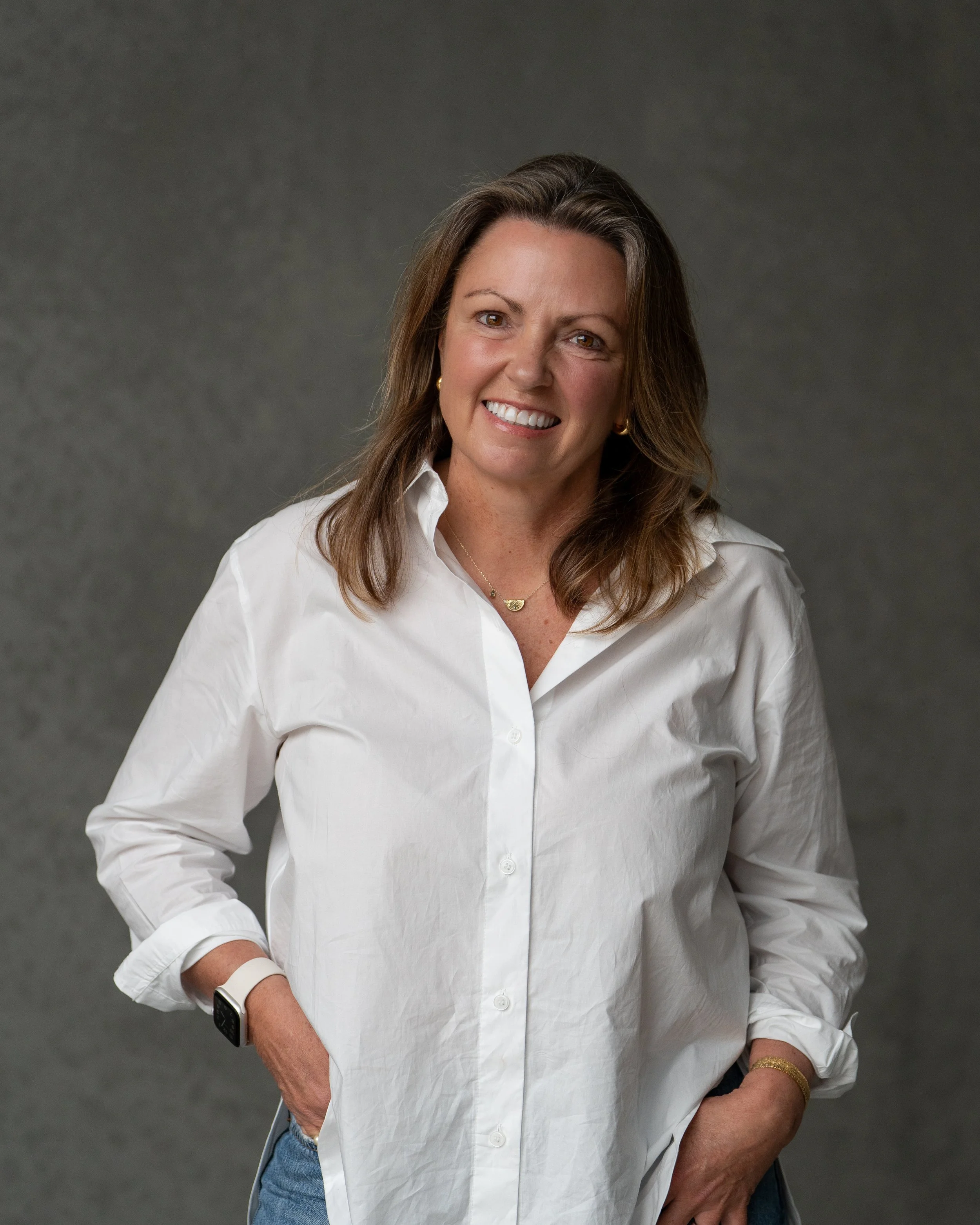 Jane Pincott, Naturopath and Nutritionist  with brown hair wearing a white button-up shirt, gold earrings, a gold bracelet, a necklace, a white watch, and blue jeans, standing against a gray background.