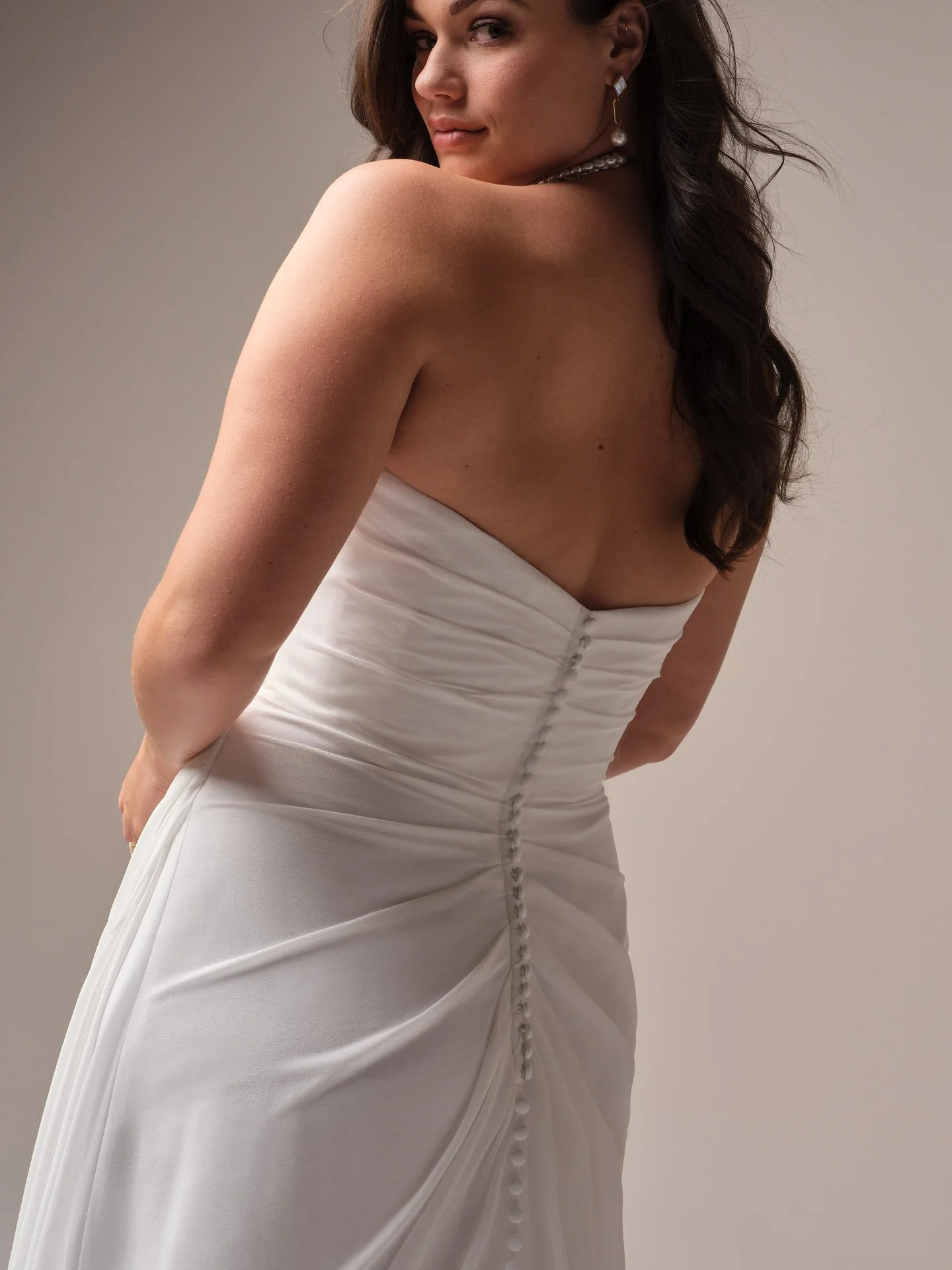 A woman in a strapless white wedding dress, looking over her shoulder, with earrings and a pearl necklace.