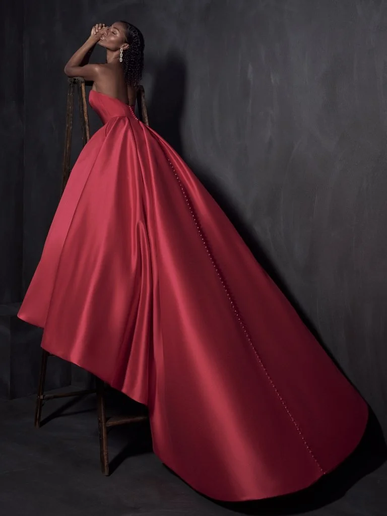 A woman in a strapless, bright red, floor-length gown with a dramatic train that creates a flowing silhouette, standing next to a dark wall.