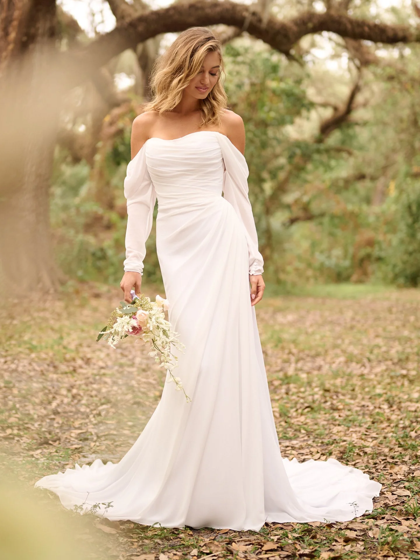 A bride in a white wedding dress holding a floral bouquet in an outdoor setting with trees and fallen leaves.