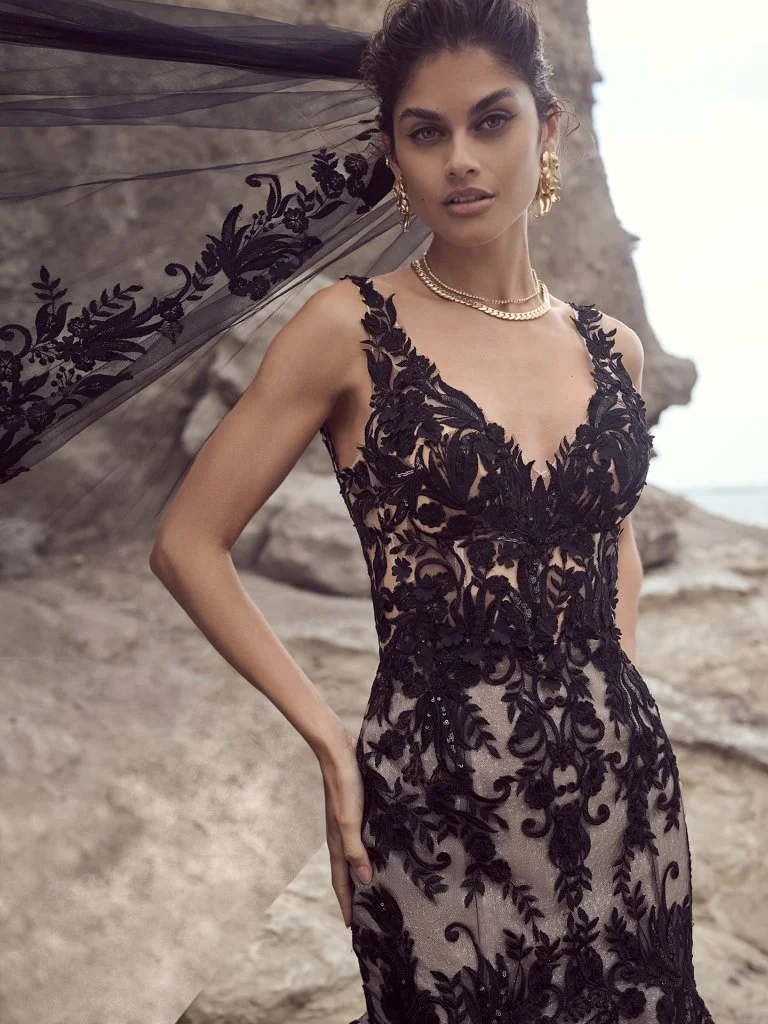 A woman wearing a black lace dress with floral embroidery and sheer fabric, standing outdoors near rocky terrain.