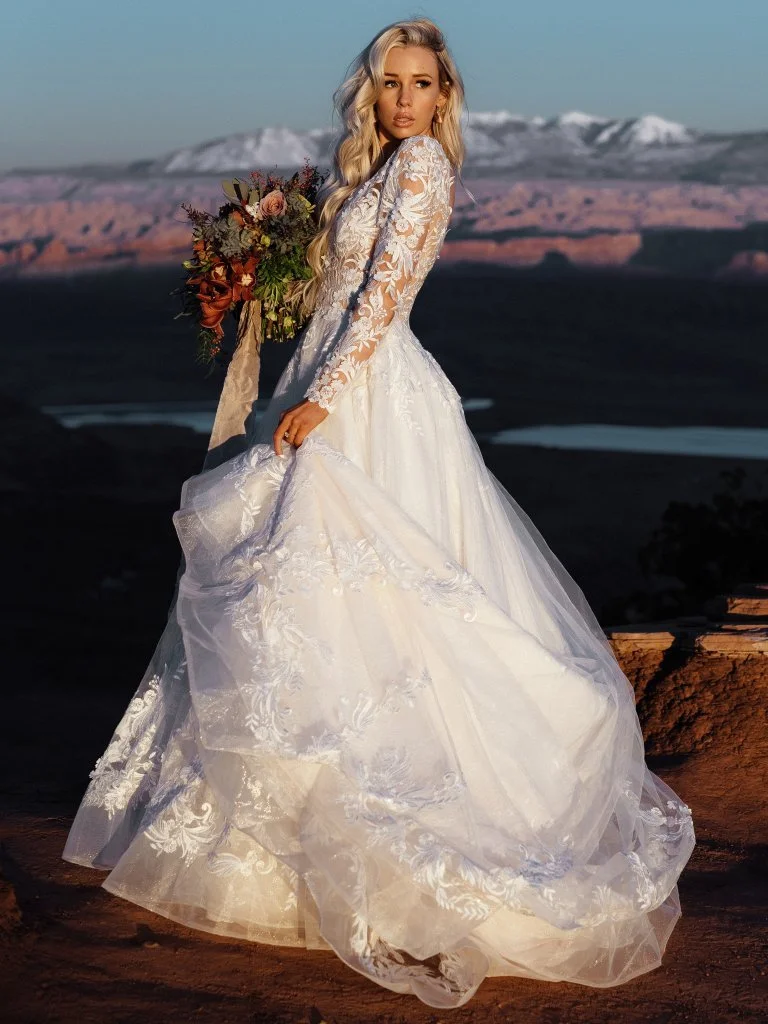 A woman in a white wedding dress holding a bouquet stands outdoors with a mountainous landscape in the background.