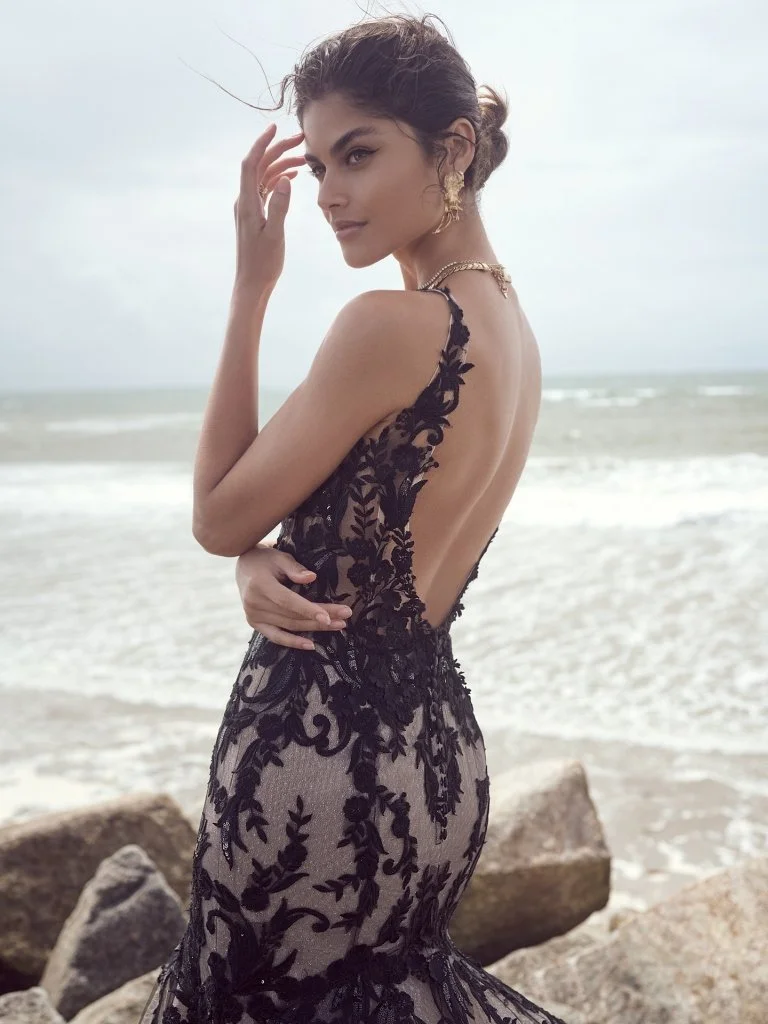 A woman in a black lace dress standing on rocks near the ocean.
