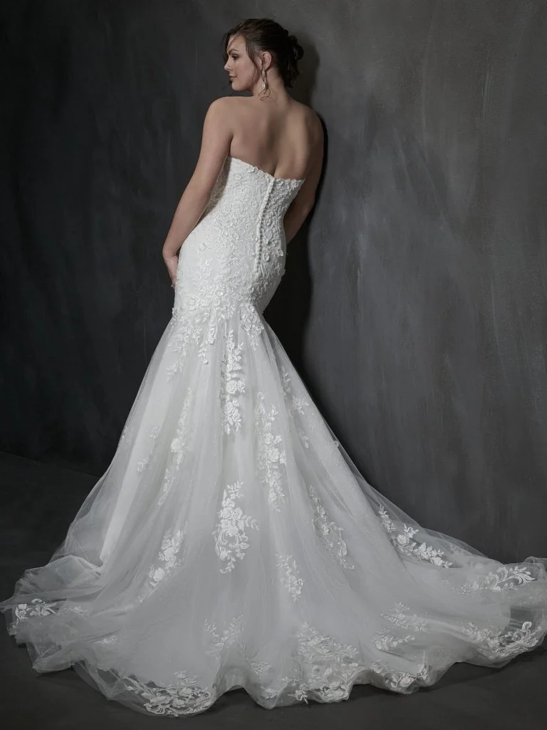 Woman in a strapless white wedding dress with lace details, standing against a dark gray wall.