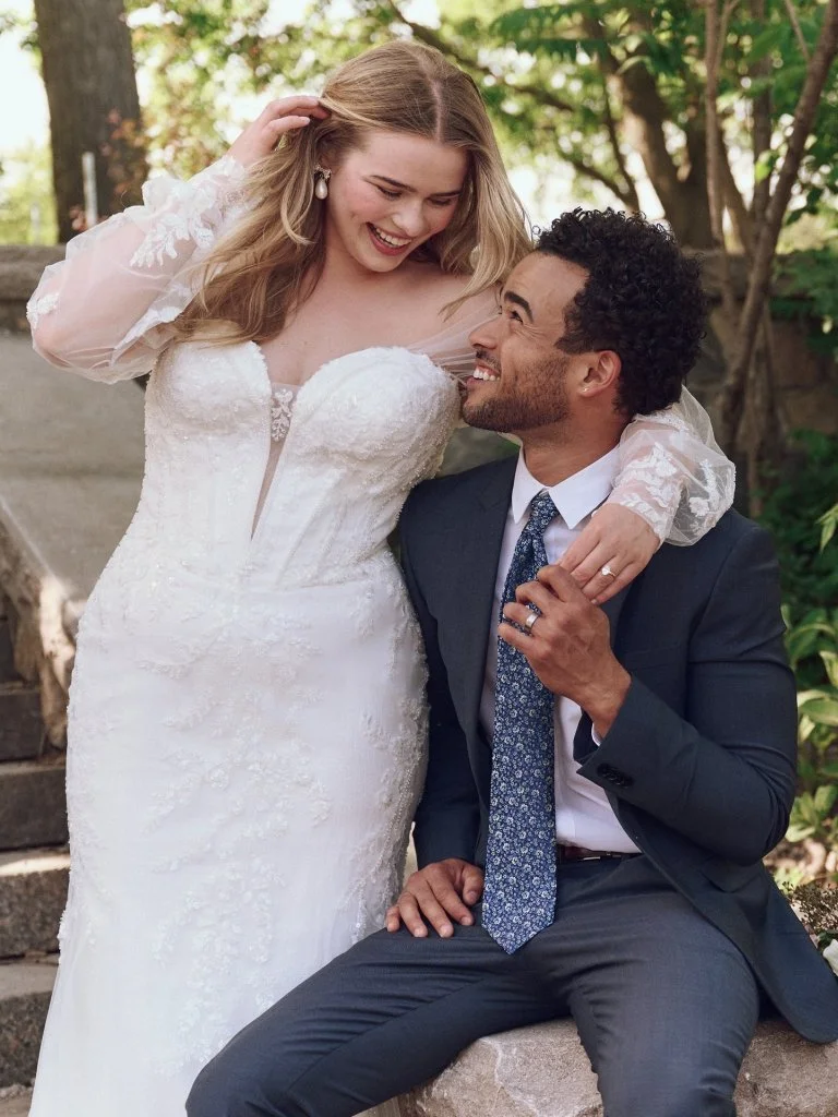 A bride and groom enjoying a moment outdoors during their wedding day, with the bride standing and the groom sitting on a stone surface, both smiling and looking at each other.