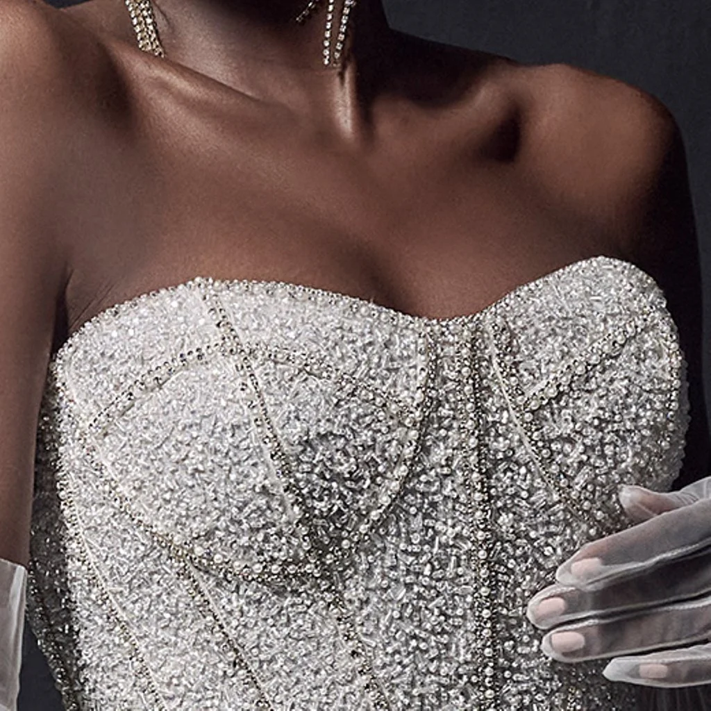 A person wearing an elaborate strapless dress covered in numerous rhinestones or crystals, with a pearl necklace, and holding a pair of white gloves.