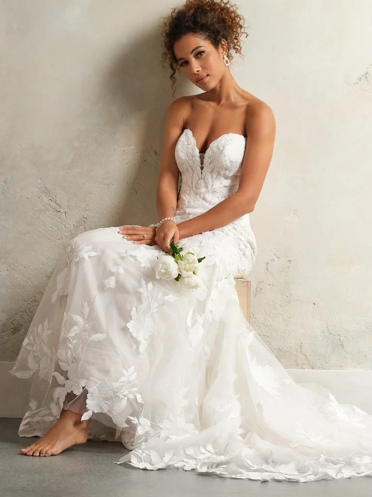 A young woman in a white lace wedding dress sitting against a light-colored wall, holding a small bouquet of white roses.