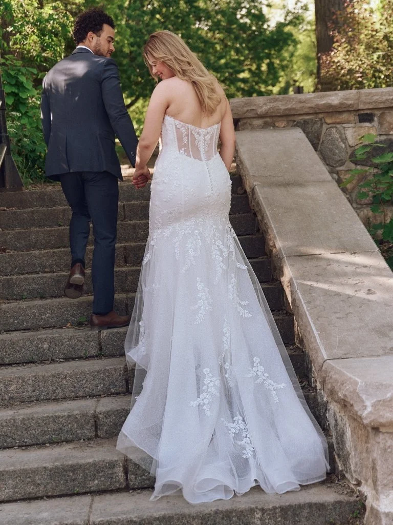 A bride and groom holding hands and walking up stone stairs outdoors, surrounded by greenery.
