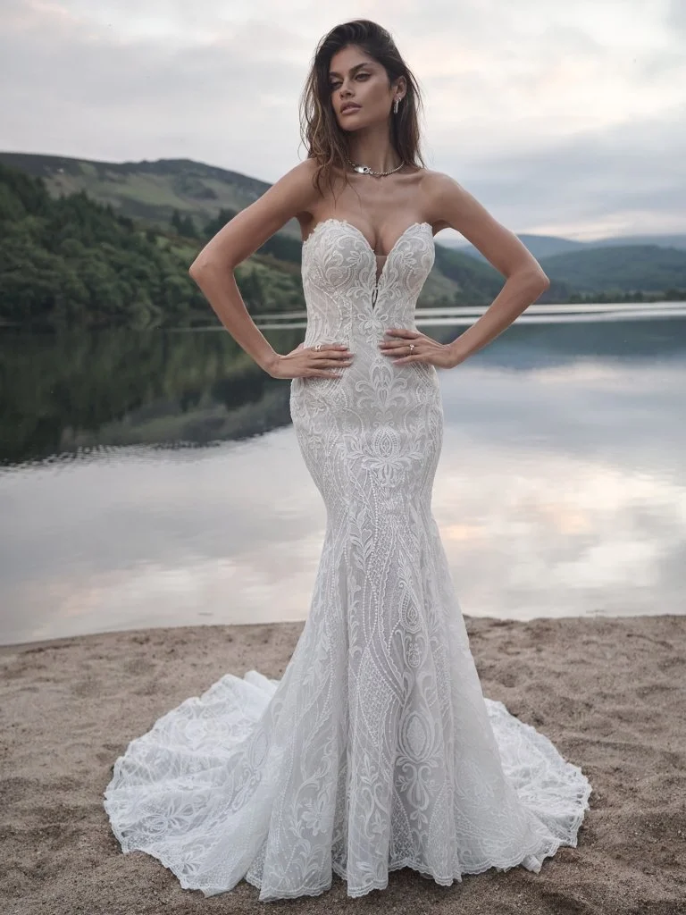 A woman in a white, strapless, lace wedding dress standing on a sandy shore near a body of water with mountains in the background.