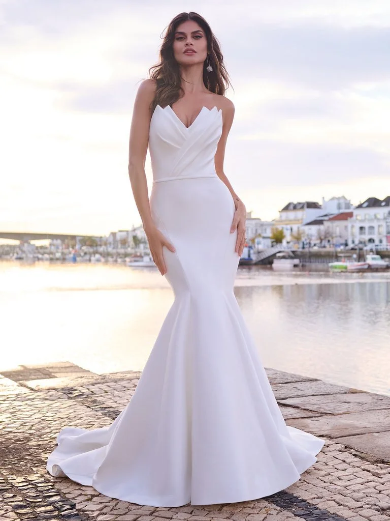 A woman in a white wedding gown standing on a cobblestone path near water with boats and colorful buildings in the background at sunset.