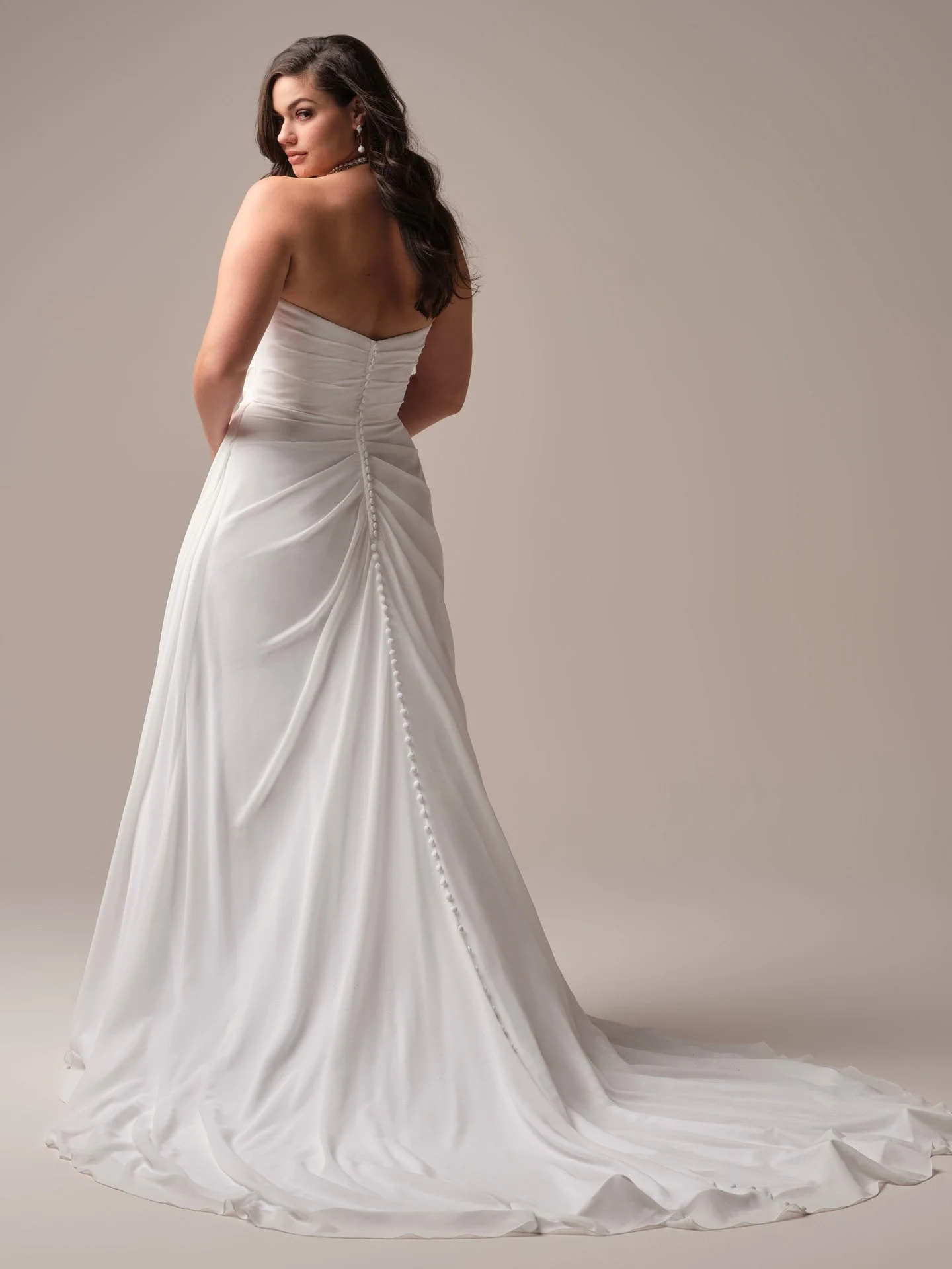 Woman in a strapless white wedding dress with ruching details, standing with her back partially turned and looking over her shoulder, against a neutral background.