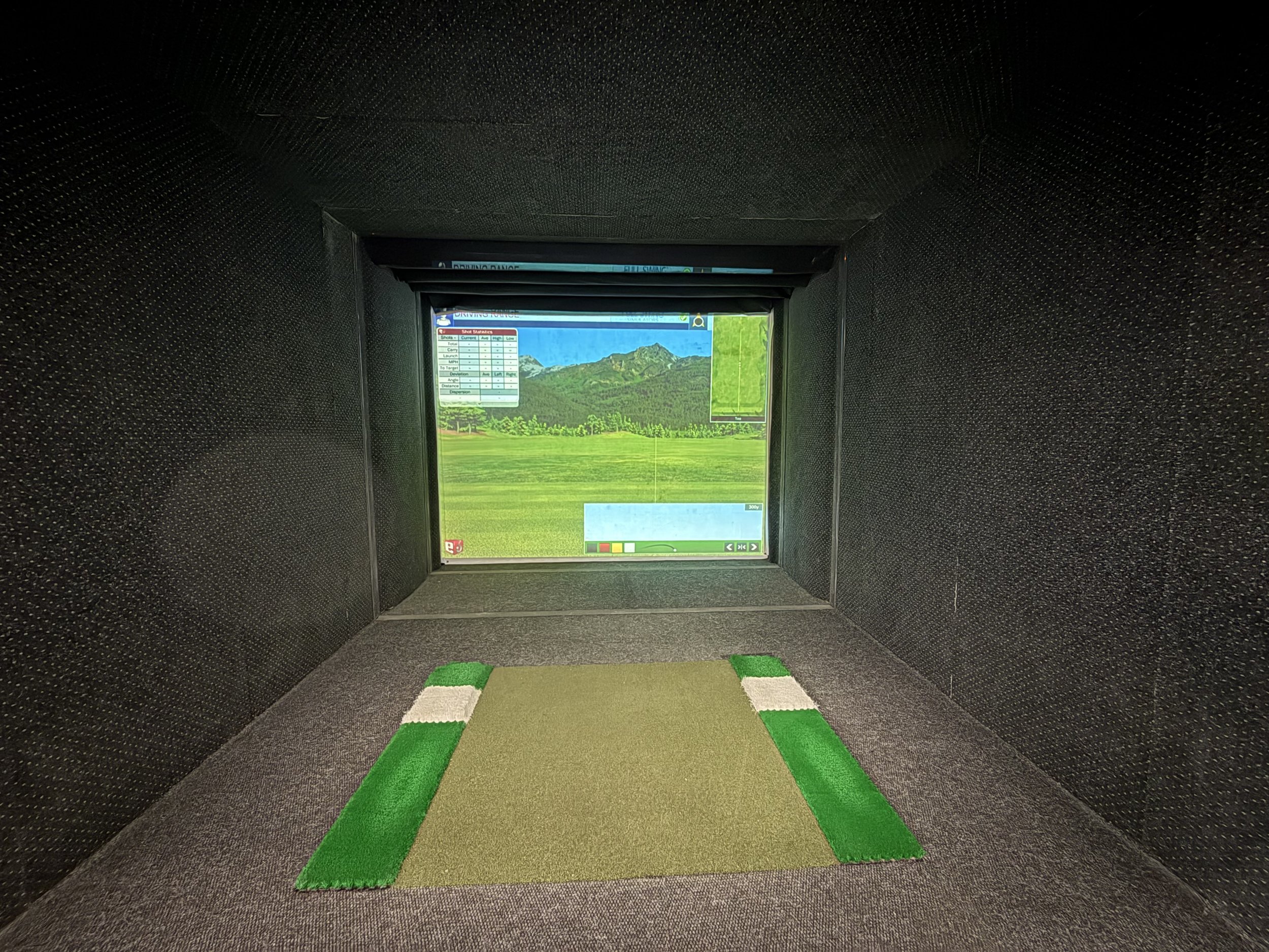 Golf simulator with a virtual golf course displayed on the screen and a small artificial golf tee mat in front.