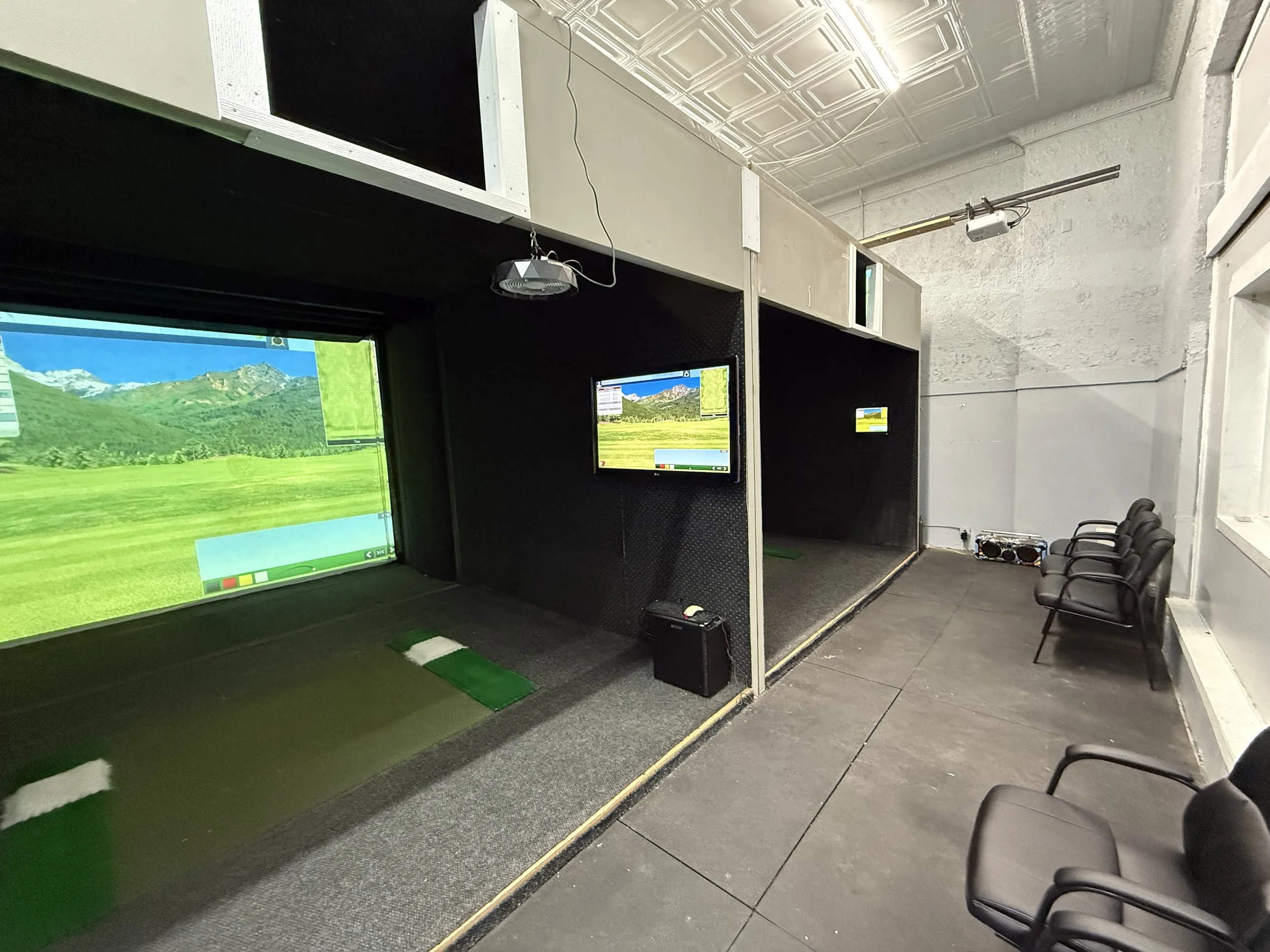 Indoor golf simulation room with large screen displaying virtual golf course, smaller monitor, four chairs, and golf simulator equipment