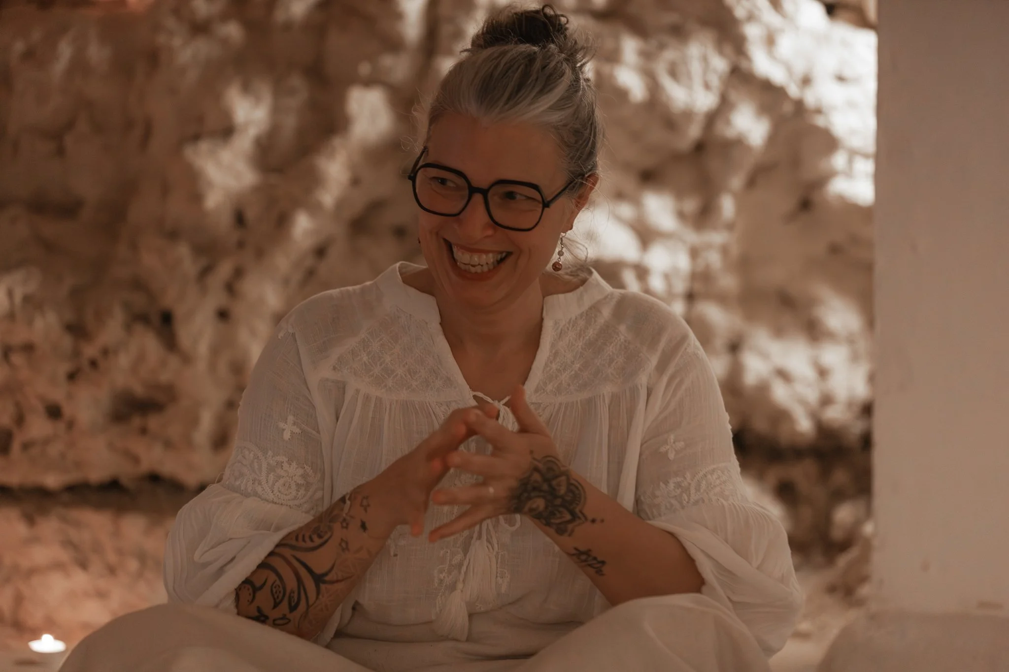 A woman with blonde hair tied back, wearing glasses and a white blouse, smiling in front of a textured stone or wall background.