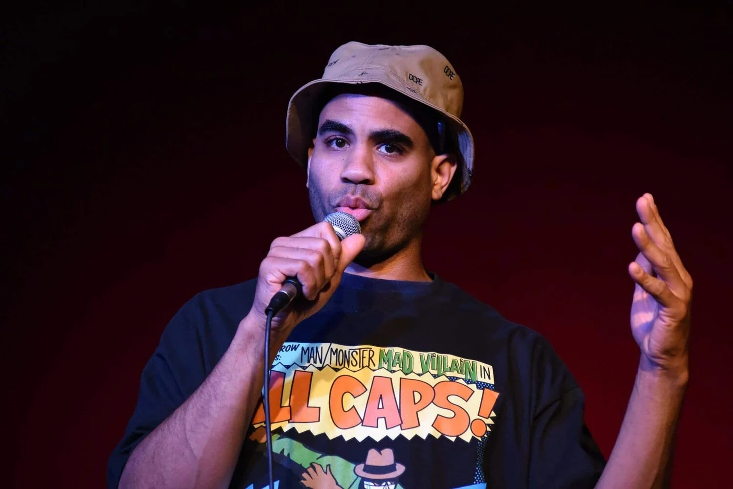 A man performing on stage, holding a microphone and wearing a beige bucket hat and a black graphic T-shirt with colorful comic book-style text.