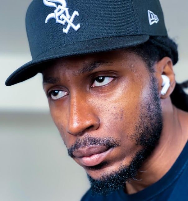 Close-up of a young man wearing a black baseball cap with white stitching and a logo, and white wireless earbuds, looking to the side with a serious expression.
