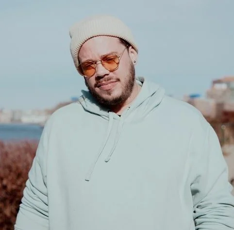 Young man with sunglasses and a beanie, wearing a light hoodie, standing outdoors near a body of water with buildings in the background.