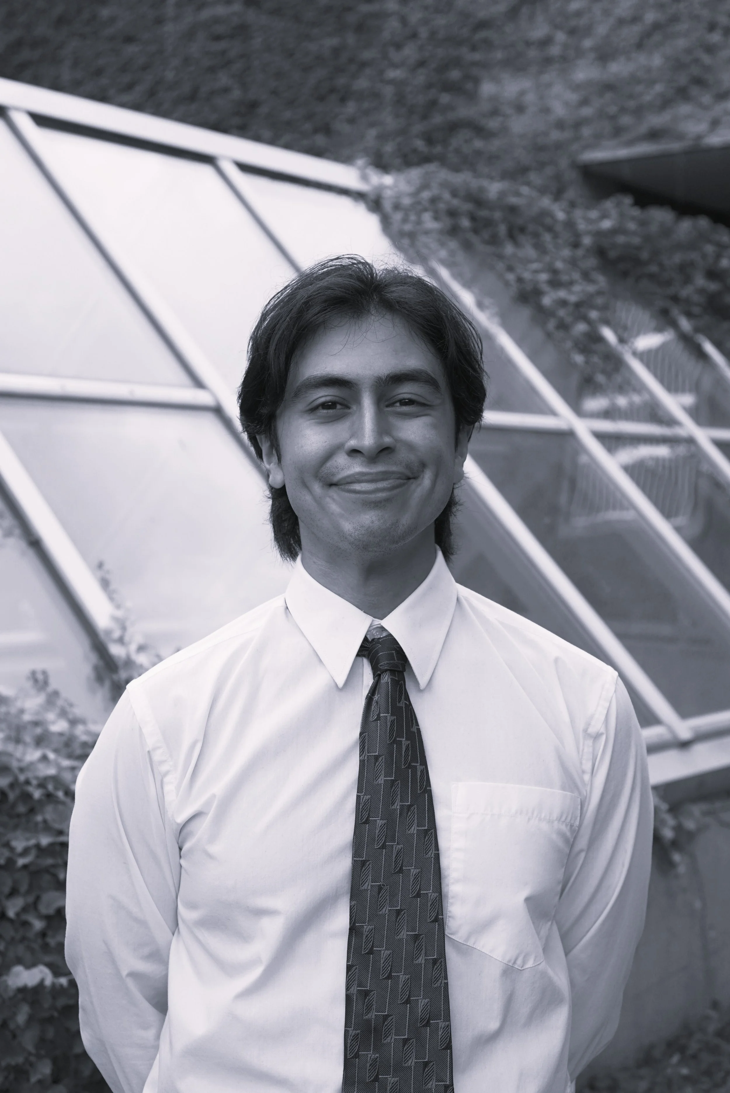 Rami is wearing a dress shirt and tie, standing infront of a greenhouse structure. He has brown hair and is smiling at the camera.