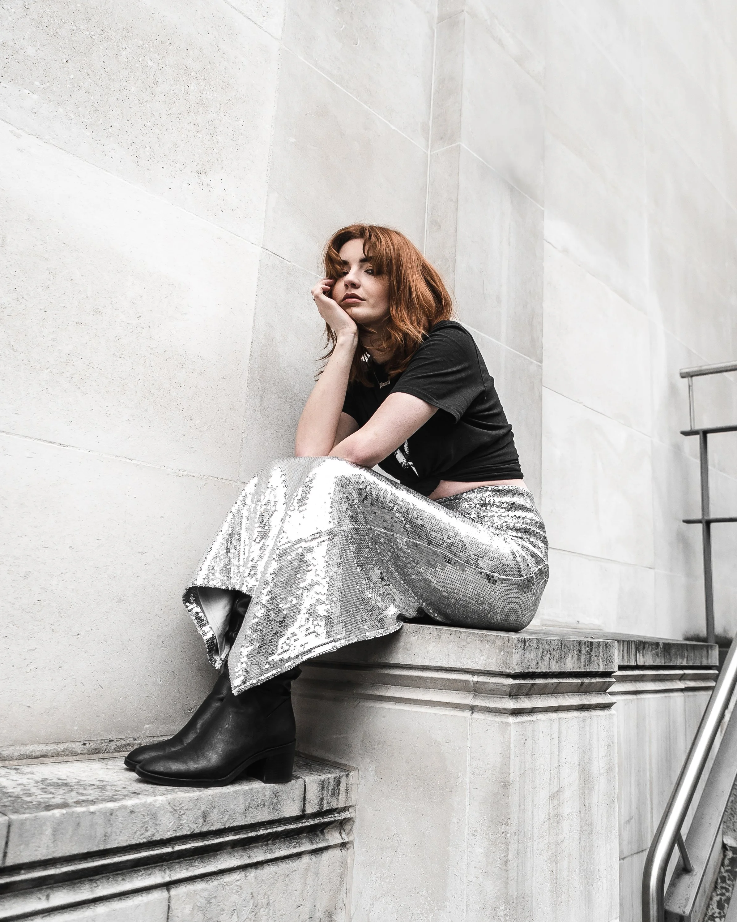 A young woman with red hair sits against a light grey stone wall, wearing a black T-shirt and shiny silver sequined pants. She rests her face on her hand, looking at the camera, with black boots visible under her pants. The setting appears to be an o