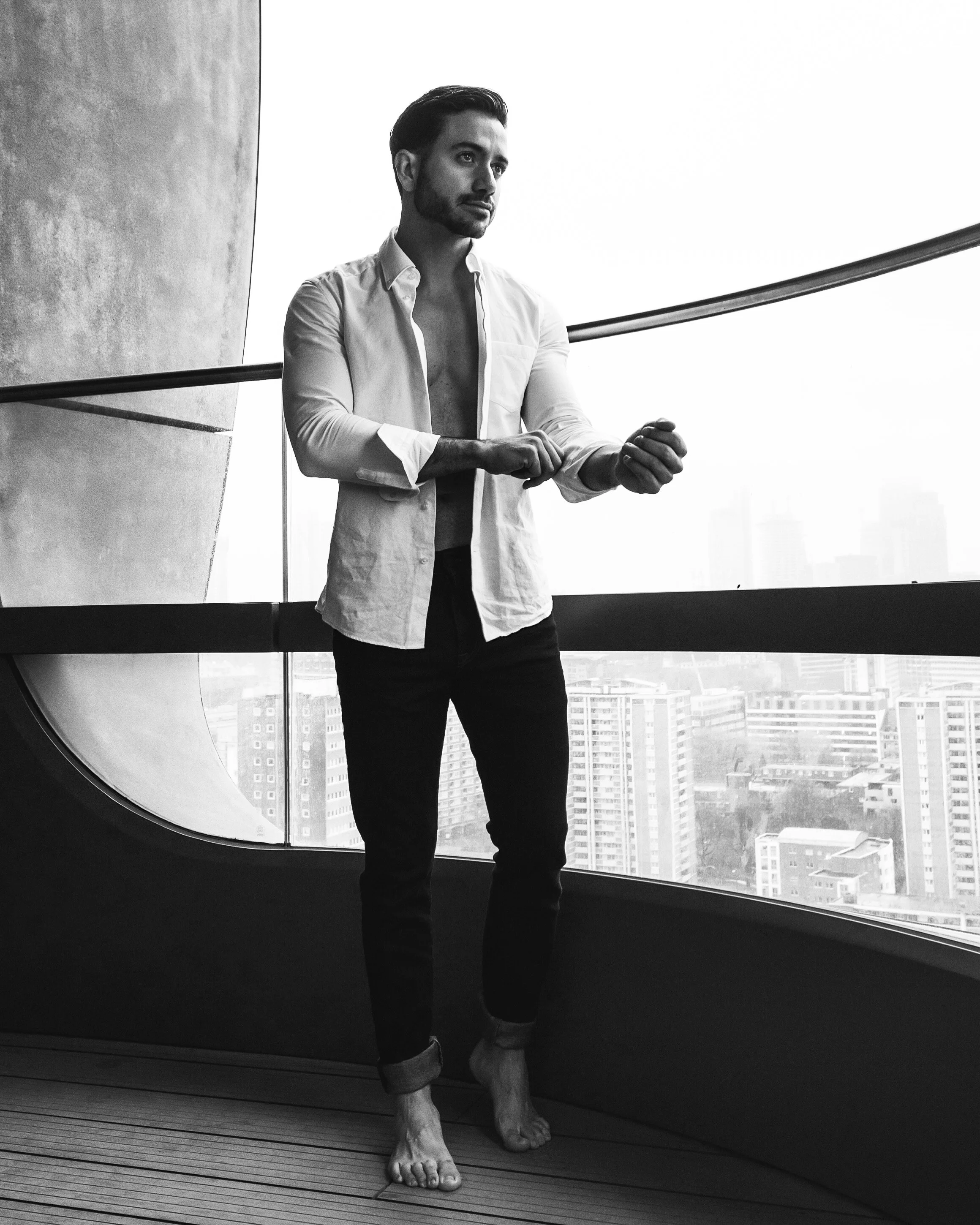 A man with a beard and mustache, wearing a partially unbuttoned white shirt and rolled-up jeans, standing barefoot near a large window in a high-rise building, with a city skyline visible in the background.