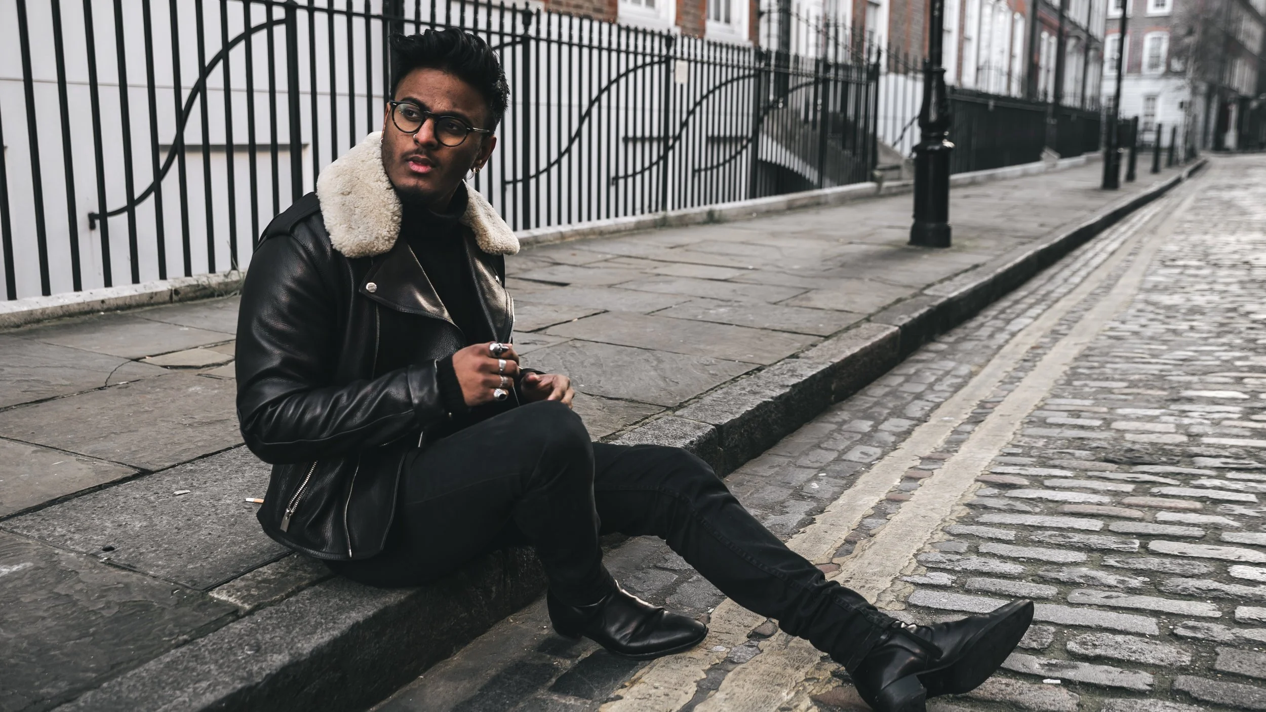 A young man with dark hair, glasses, and earrings sitting on the sidewalk against a curb, wearing a black leather jacket with a shearling collar, black pants, and black boots, with a black metal fence and brick buildings in the background.