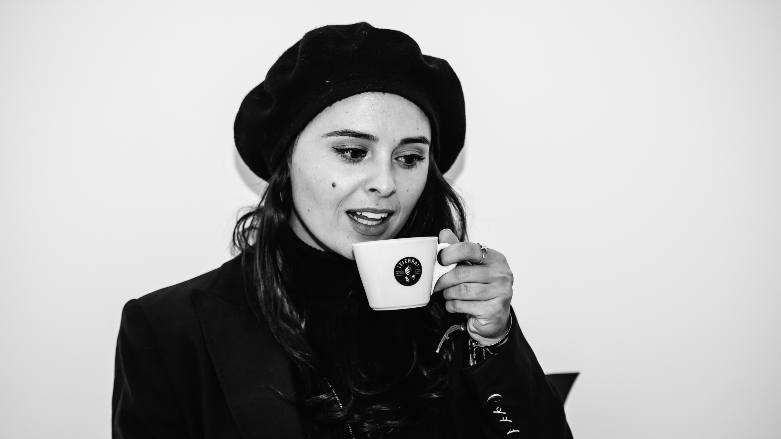 A young woman with dark hair and a mole on her cheek, wearing a black beret and black jacket, is holding a white coffee cup with a logo and looking down at it.