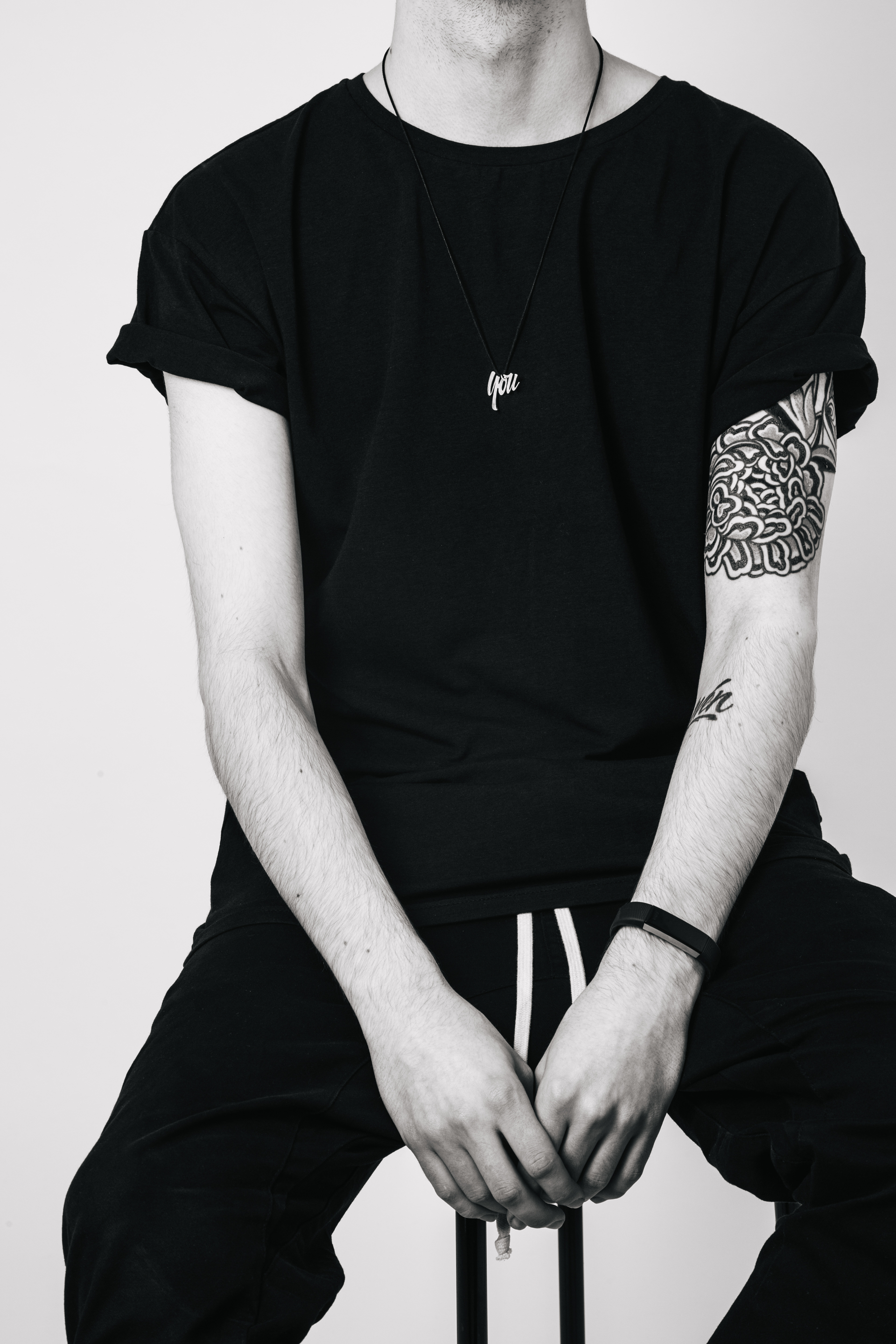 A black-and-white photo of a man wearing a black t-shirt with rolled sleeves, black pants, and a necklace with the word "you". He has tattoos on his right arm and is sitting on a black stool with his hands clasped in front of him, against a plain light background.