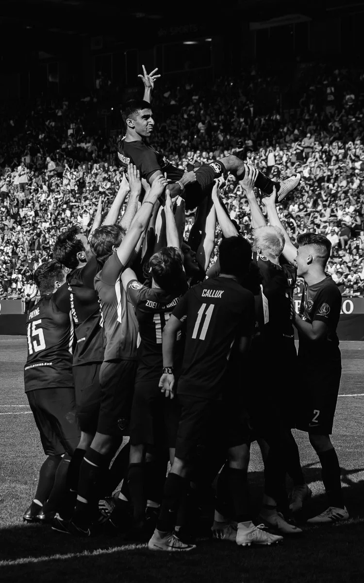 Soccer team celebrating by lifting a player onto their arms on a soccer field with a large crowd in the background, black and white photo.