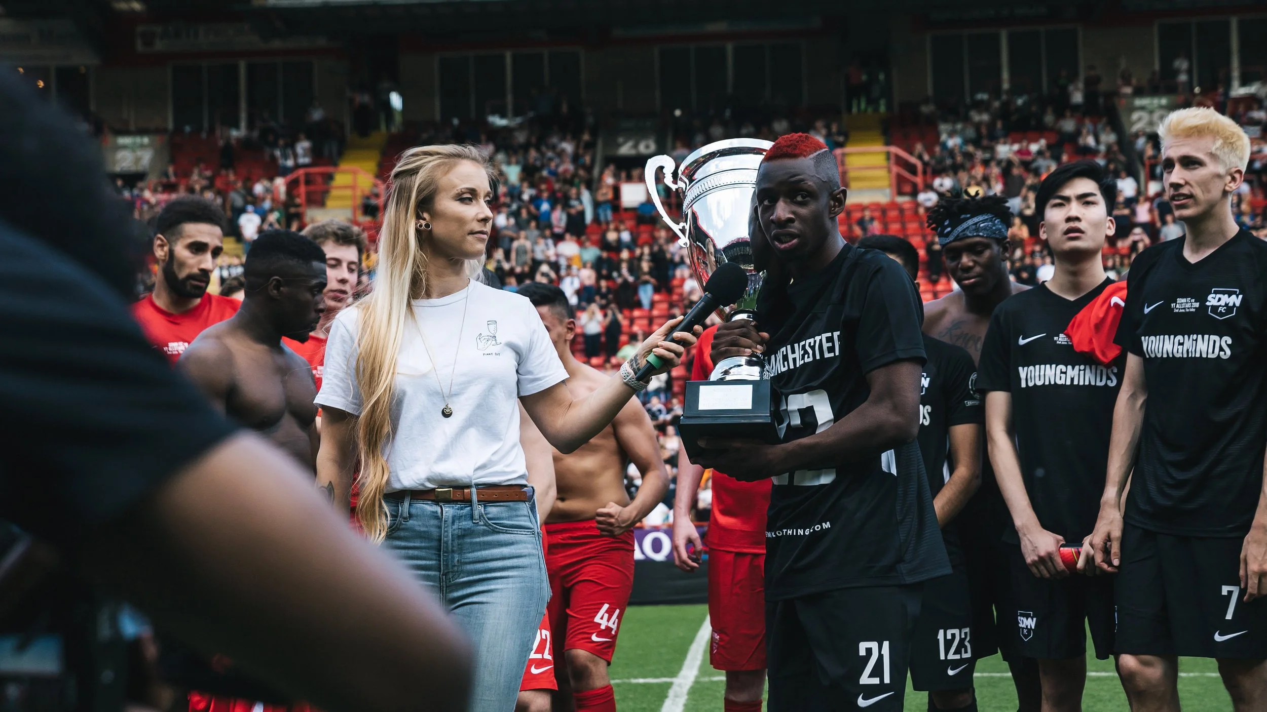 A woman with long blonde hair interviewing a soccer player holding a trophy on a soccer field. Several other players are standing around, and the stadium is filled with spectators in the background.