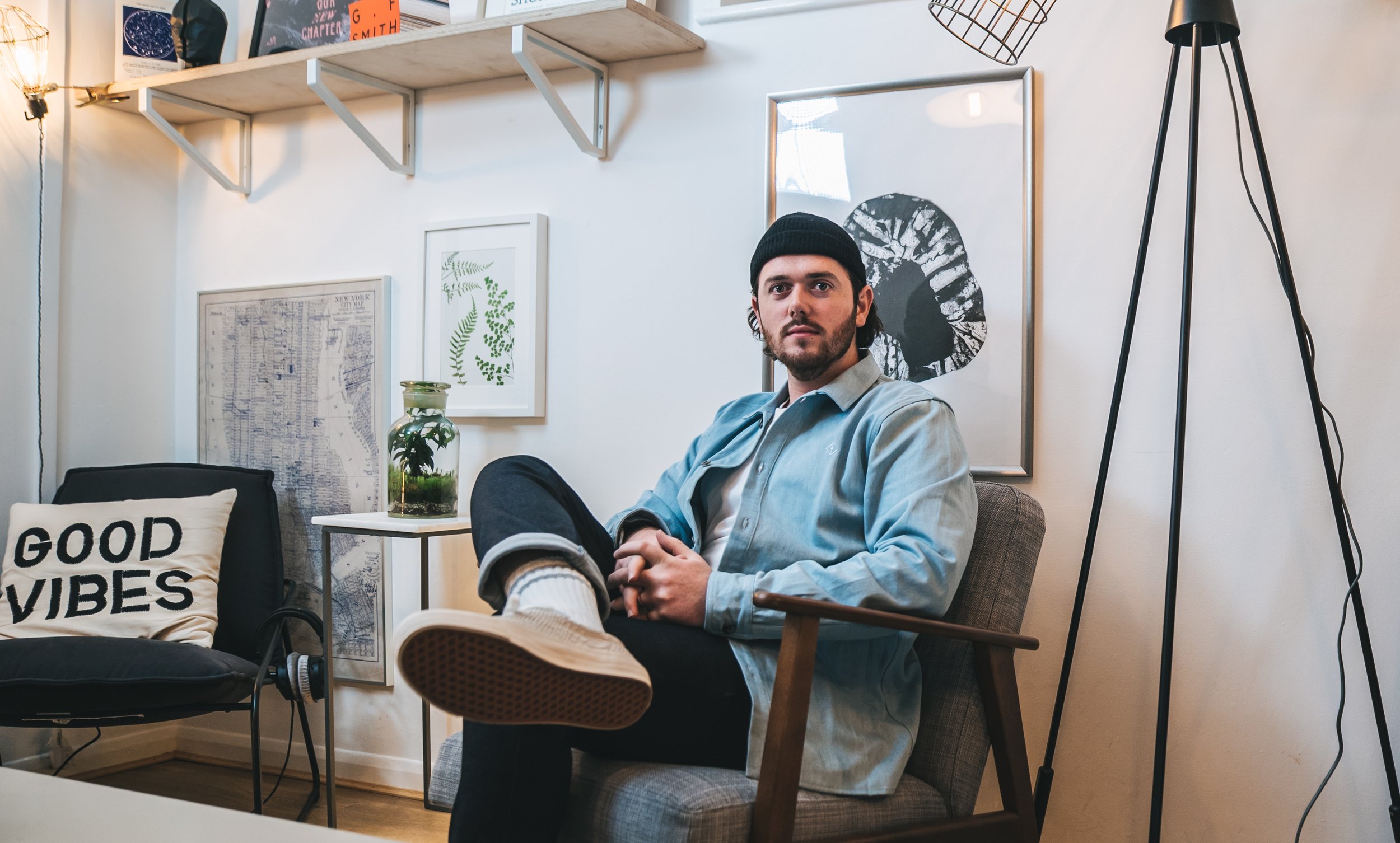 A man with dark hair and a beard wearing a black beanie, light blue shirt, and jeans, sitting in a modern living room with white walls, framed artwork, a lamp, and a pillow reading "Good Vibes."