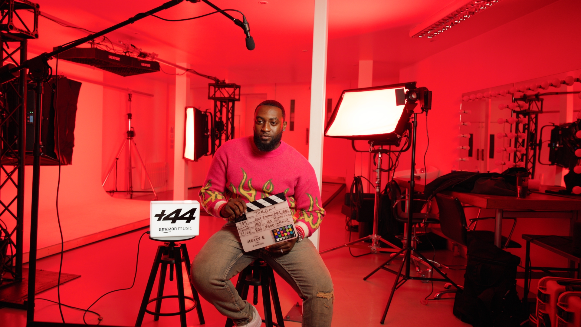 A man sitting on a stool holding a clapperboard in a professional videography studio with red lighting, surrounded by studio lights, equipment, and a backdrop.