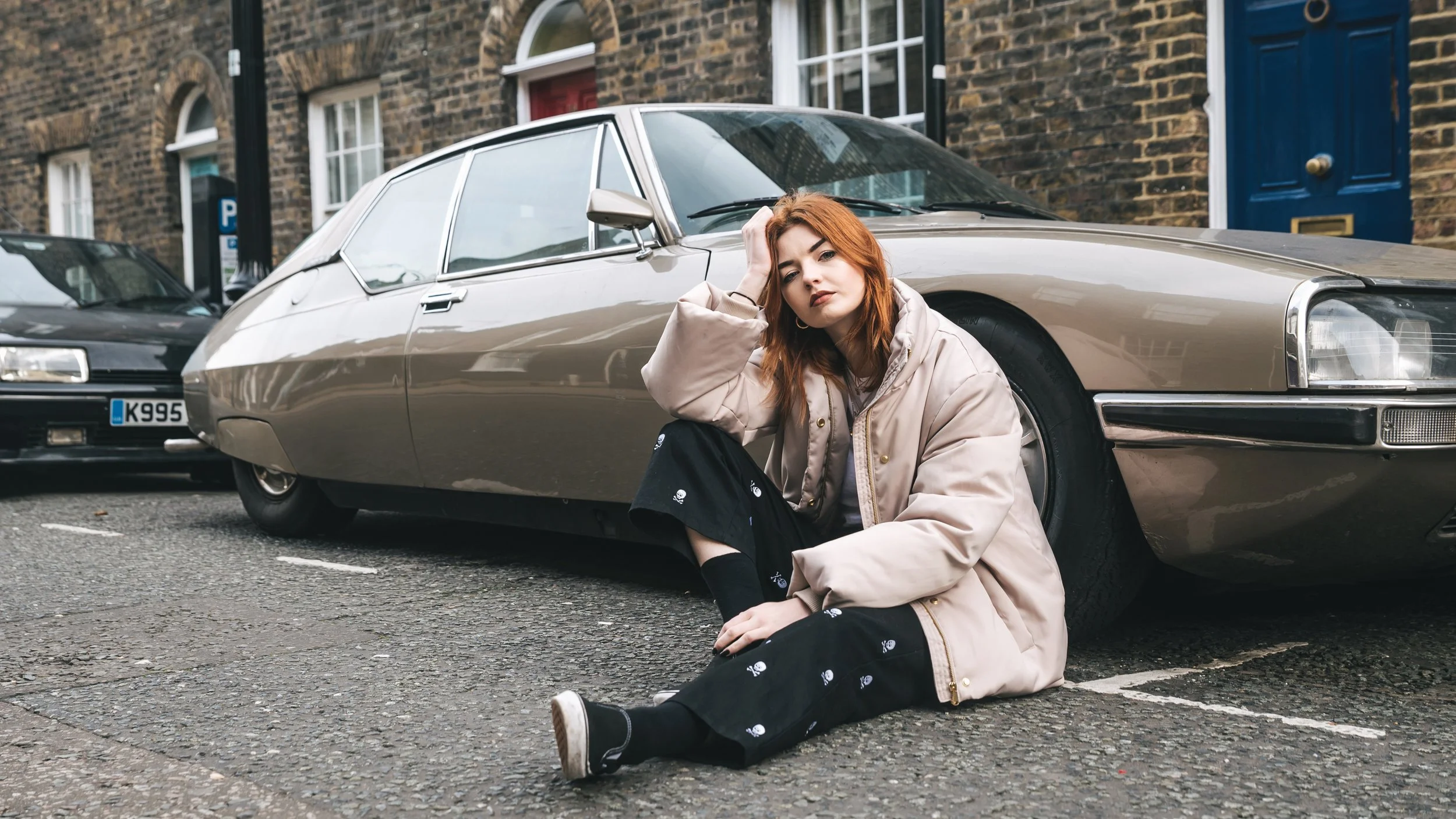 A young woman with red hair sitting on the street in front of a vintage silver car, resting her head on her hand and looking at the camera.