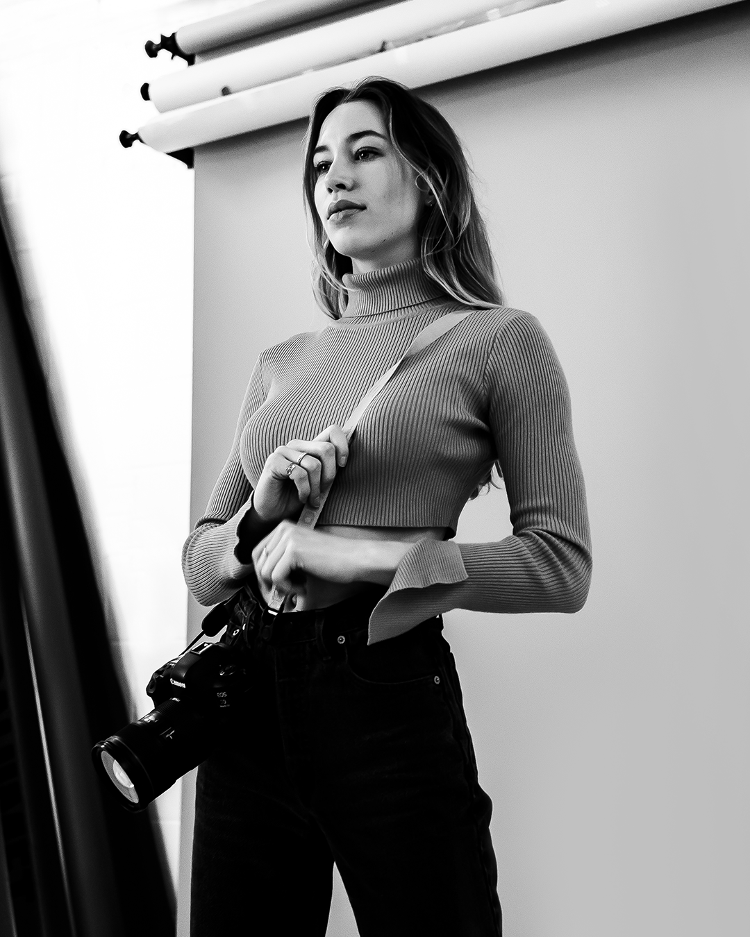 A woman with shoulder-length hair in a studio setting, holding a camera with a strap. She is wearing a ribbed turtleneck sweater and high-waisted pants, posed in front of a backdrop roll.