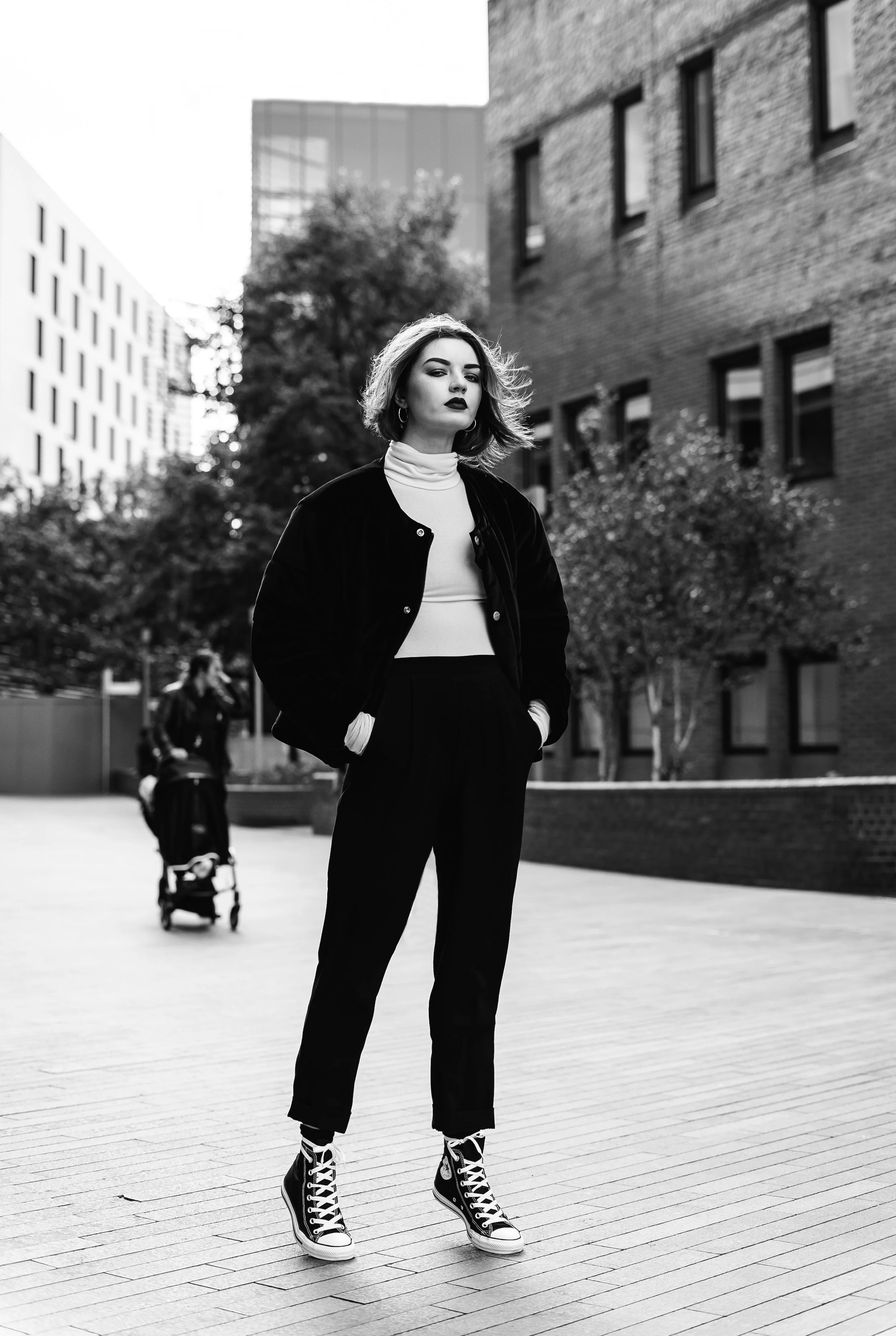 A young woman with medium-length hair and dark lipstick stands confidently on a city sidewalk, wearing a black jacket, white turtleneck, black pants, and high-top sneakers. In the background, there are buildings, trees, and a person with a stroller.
