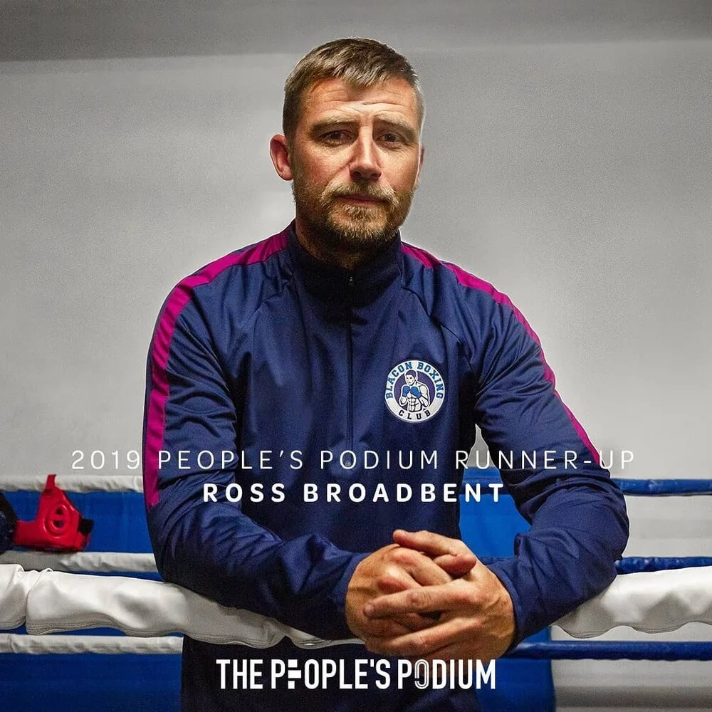 A man with short hair and a beard standing in a boxing gym, wearing a blue sports jacket with pink accents and a club logo, with boxing rings and equipment in the background.