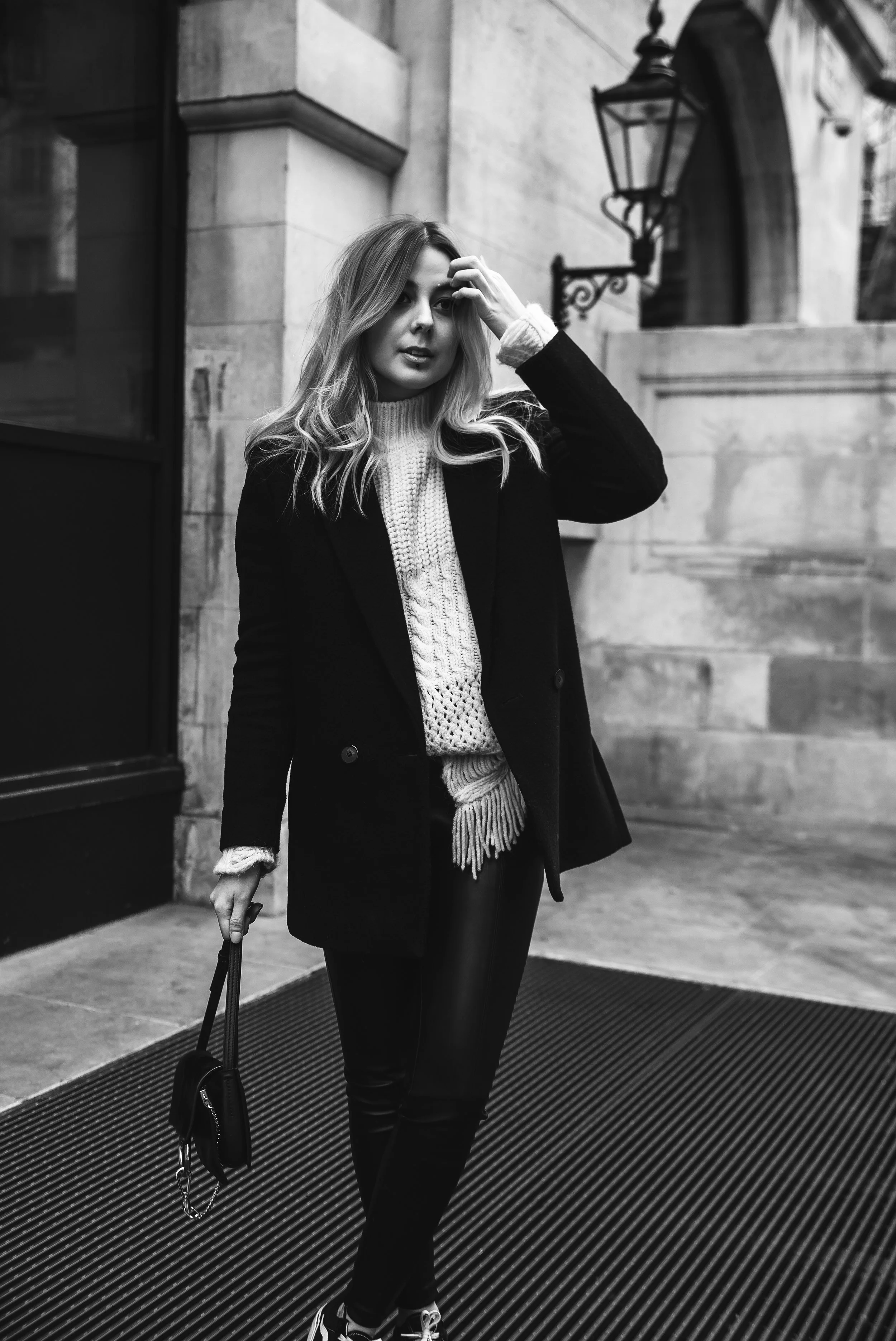 A woman with wavy, shoulder-length hair stands outside a building with stone walls and a lantern on the wall. She is wearing a knit sweater, a dark coat, and dark pants. She holds a handbag in her right hand and is touching her hair with her left han