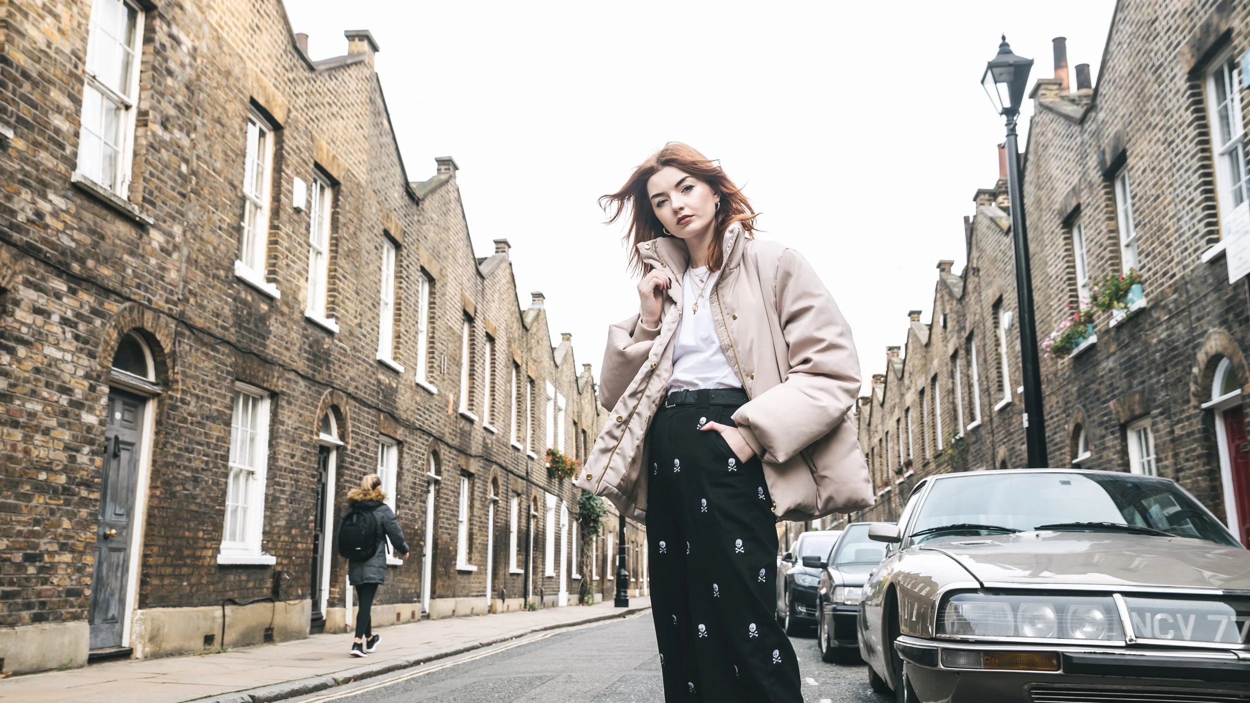 A young woman with reddish hair stands on a city street with brick buildings and parked cars, wearing a beige jacket and black pants with skull and crossbones pattern.