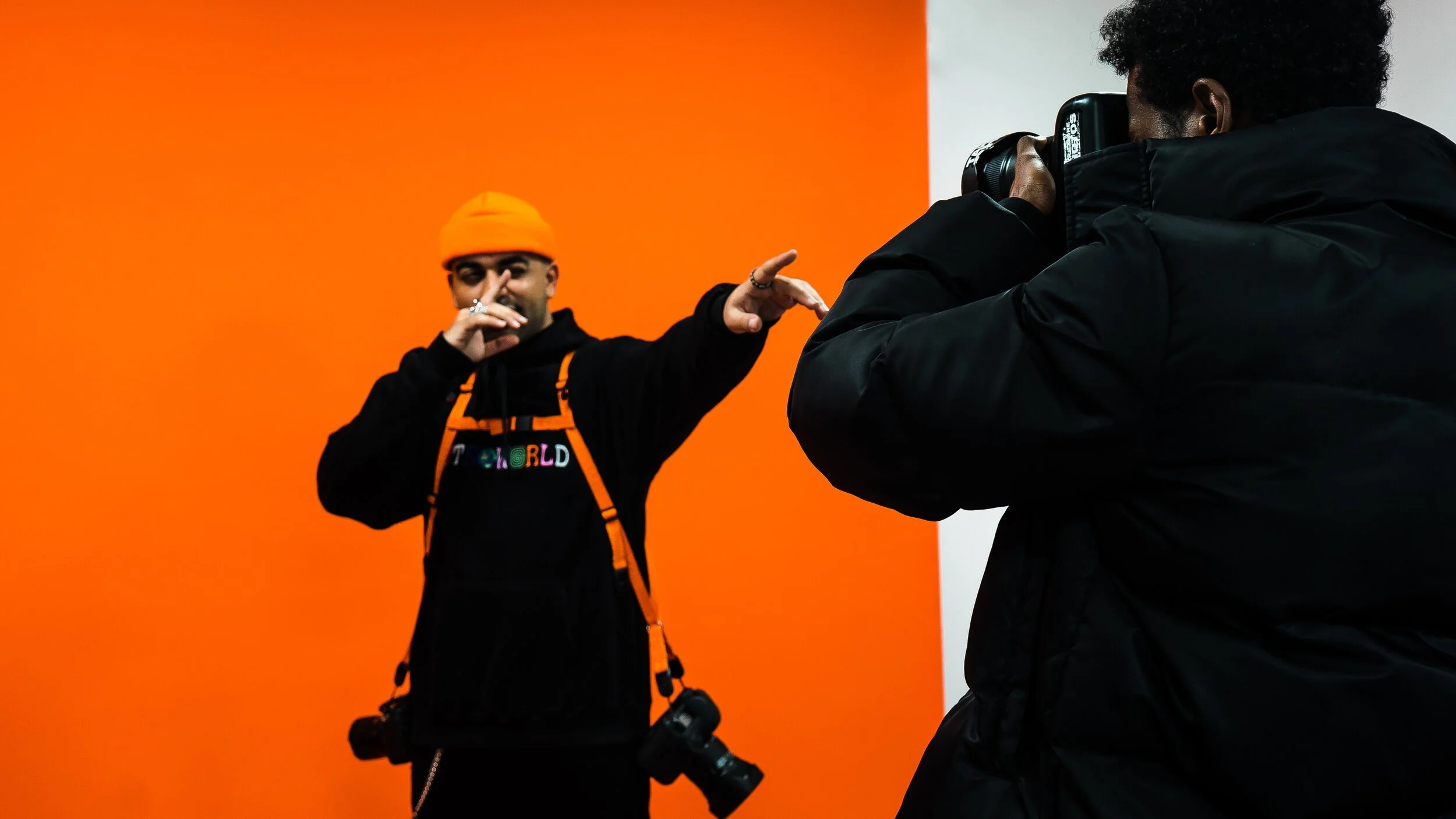 Two photographers working in a studio with a vivid orange background. One is pointing while the other is taking a photo.