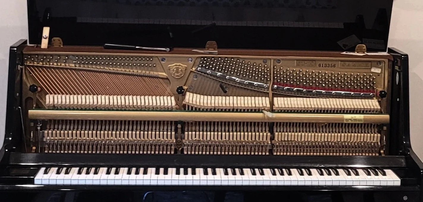 A close-up view of the inside of an open upright piano, showing the cast iron plate, tuning pins, strings, and hammers above the white and black keyboard keys.