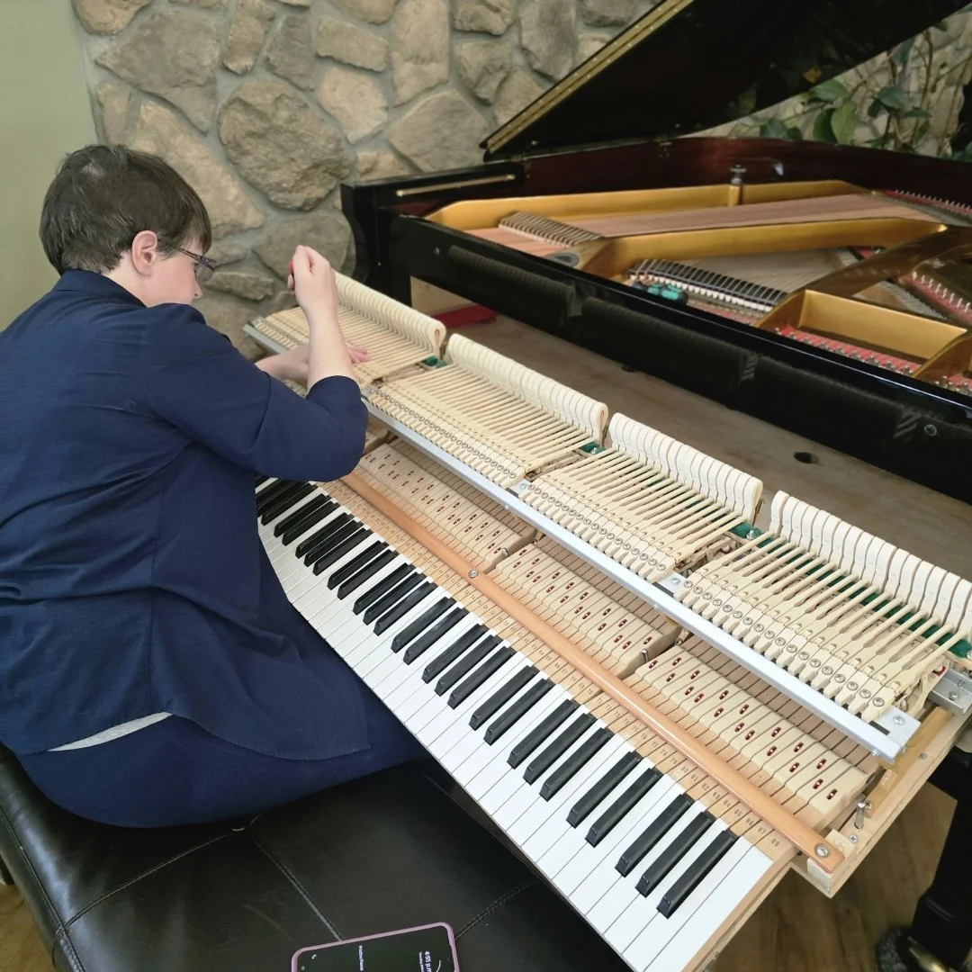 Audi Lou Nordby, Saskatoon piano tuner, sits on a piano bench, repairing the inner mechanics of a grand piano with the lid open, revealing the hammers and strings.