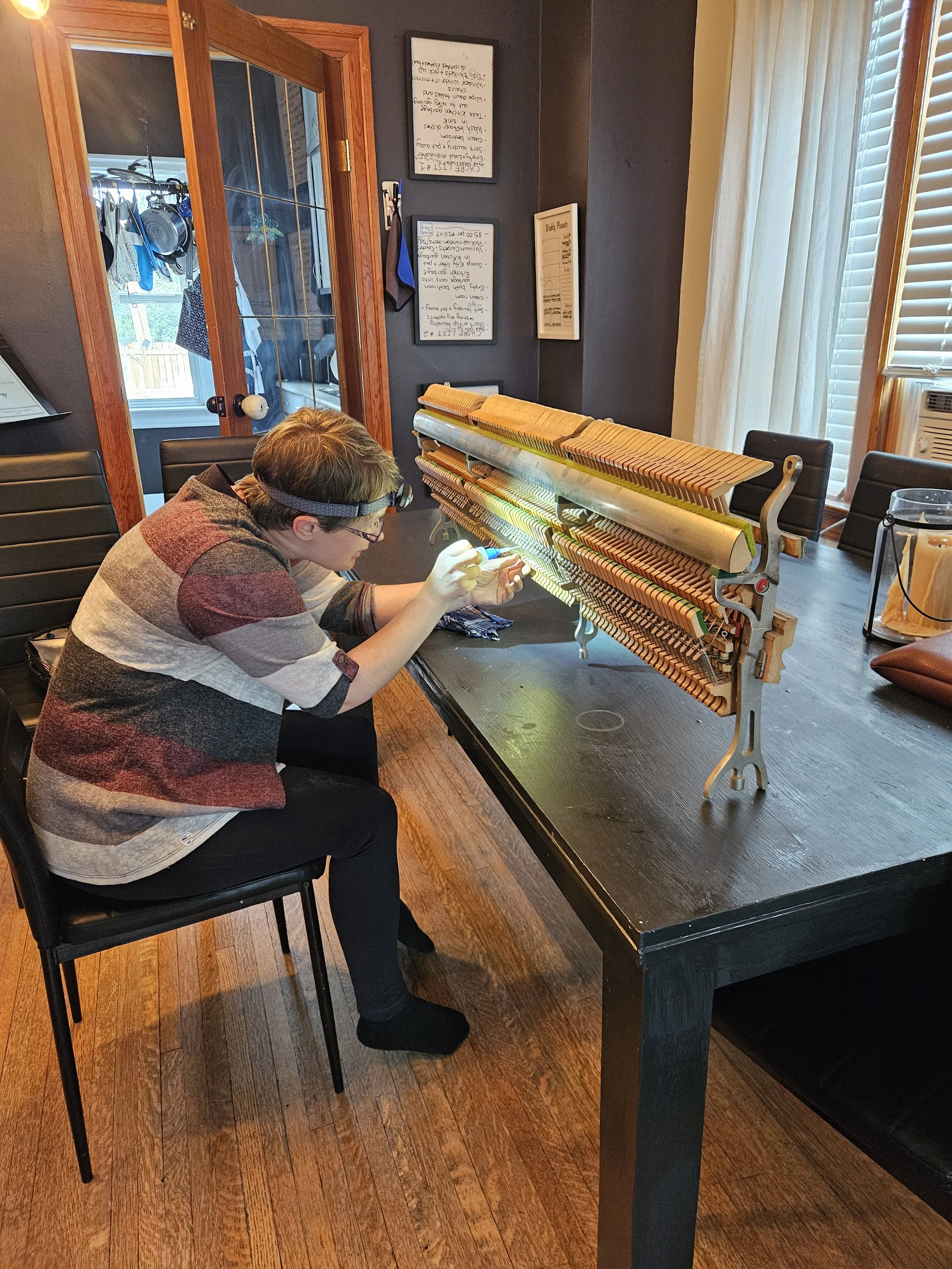 Registered Piano Technician Audi Lou Nordby sits at a black table wearing a headlamp, carefully repairing the exposed action of an upright piano in a Saskatchewan home.