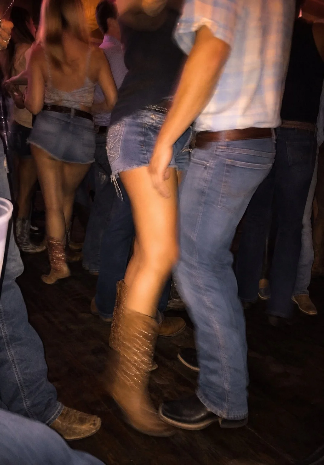 People dancing in a dimly lit setting, wearing casual and country-style clothing, including denim shorts and cowboy boots.