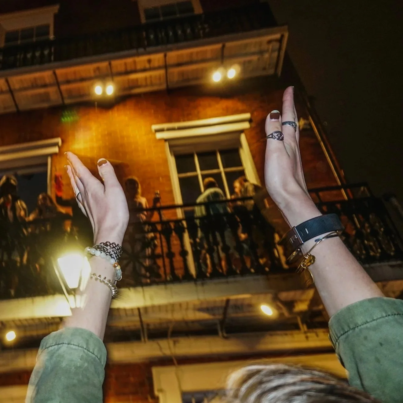 Person with jewelry and rings holding their arms up in front of an outdoor balcony at night with people gathered on the balcony, illuminated by warm outdoor lighting, in front of a brick building with windows.