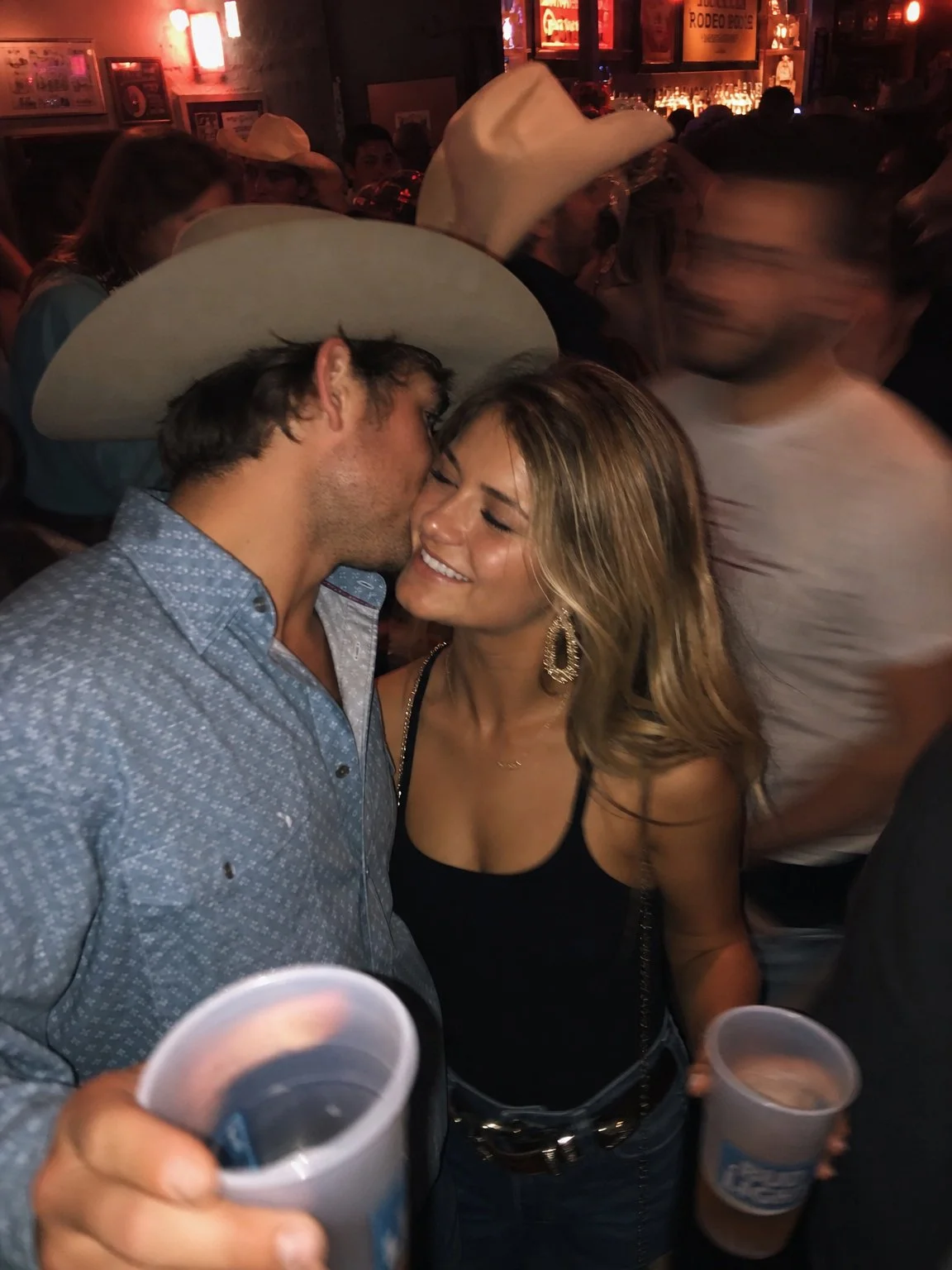 A man wearing a large white cowboy hat and a blue shirt is kissing a smiling woman with wavy blonde hair, wearing earrings and a black top, in a crowded bar or party setting, both holding cups.